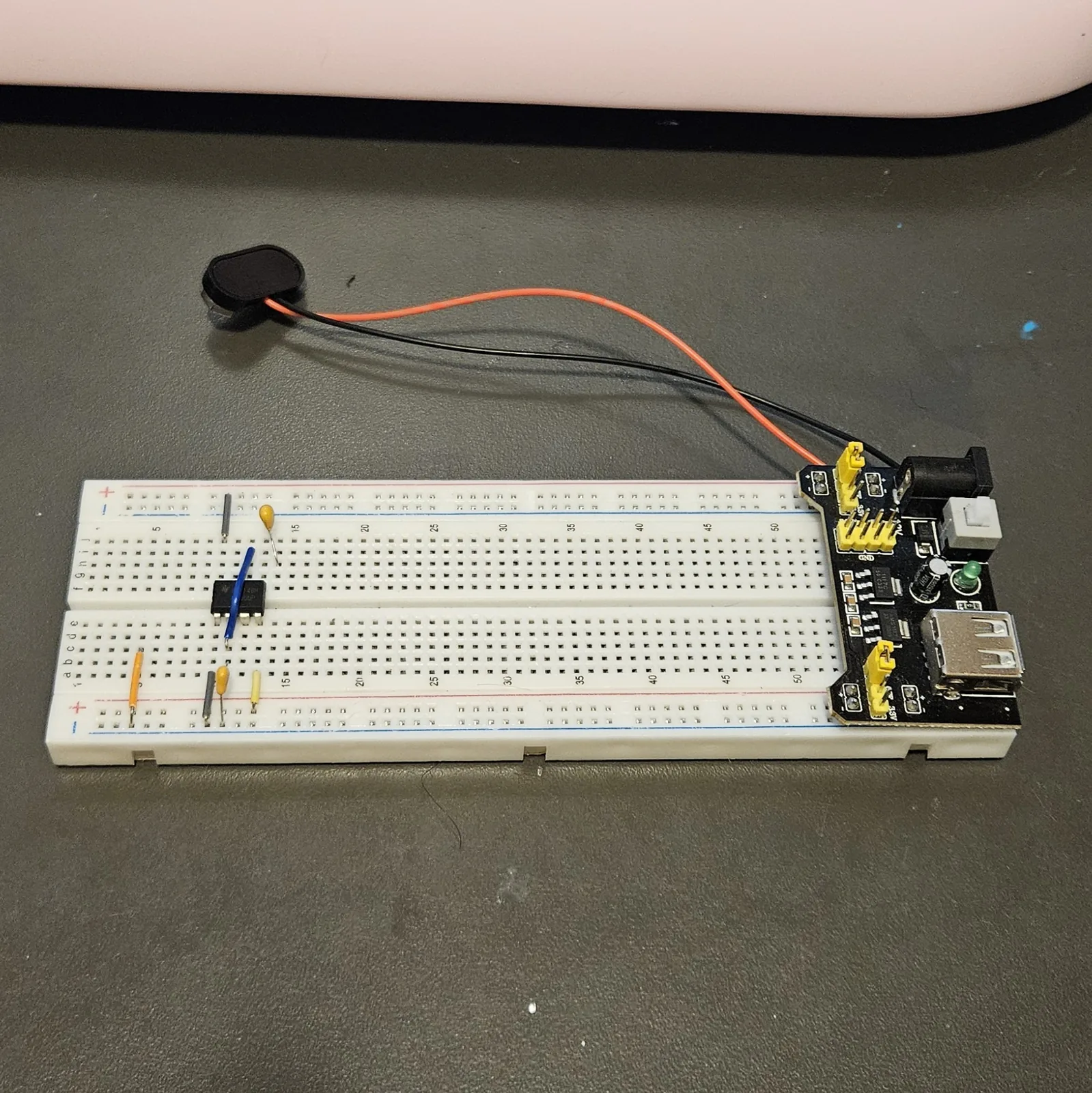 The breadboard with the small ceramic capacitors added. A yellow disc capacitor leans up toward the top rails just right of the chip and a second sits in the lower bank below it, with the blue jumper still arcing across the timer. The power module and 9-volt battery sit at the right and the speaker lies off the top of the board.