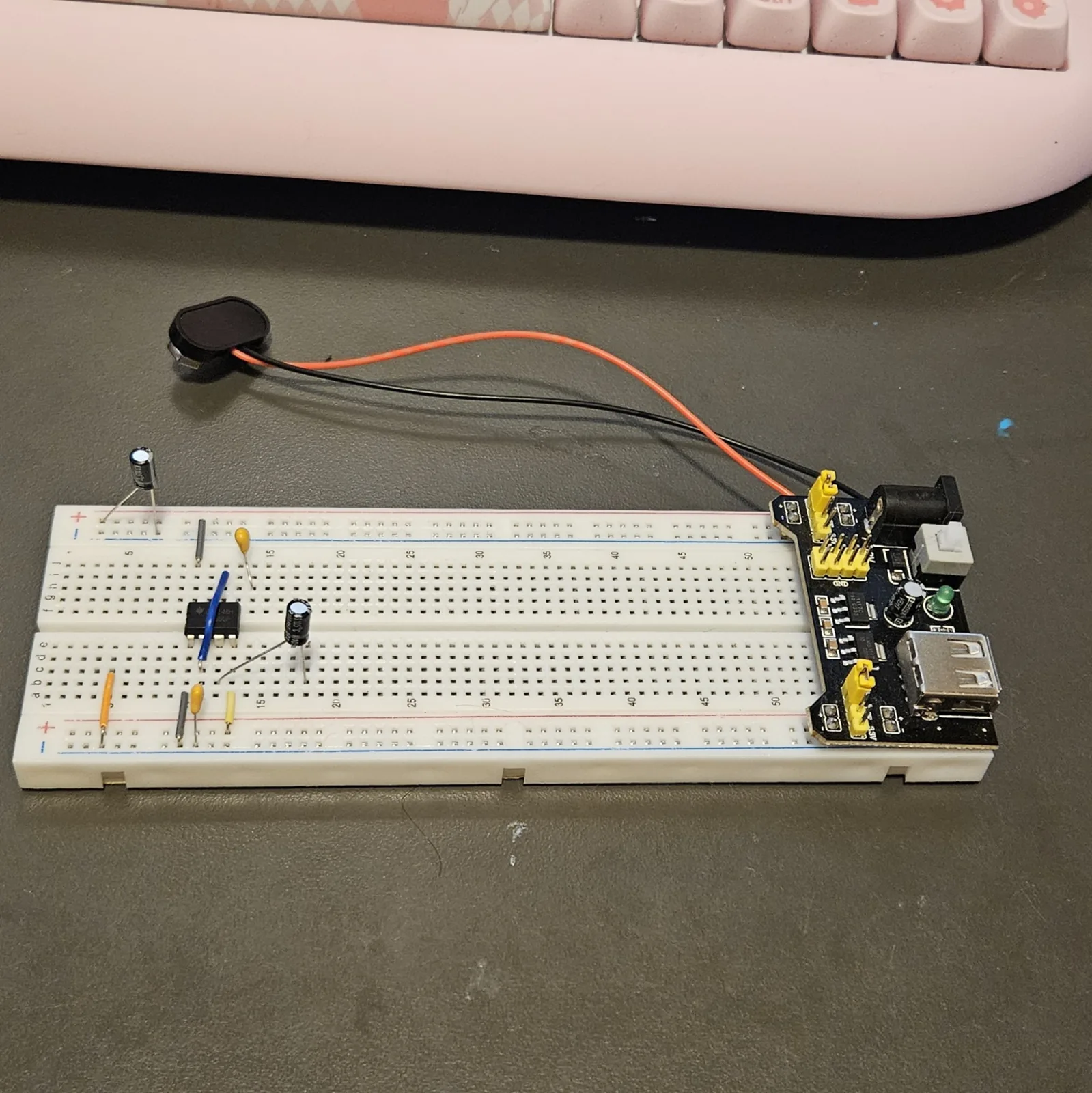 The breadboard with both electrolytic capacitors added. One black can stands at the far left with its legs in the two top rails, and a second sits in the lower bank just right of the timer chip. A gray jumper and a yellow ceramic capacitor stand between them, and the blue jumper still crosses the chip. The power module is plugged into the right-hand end.