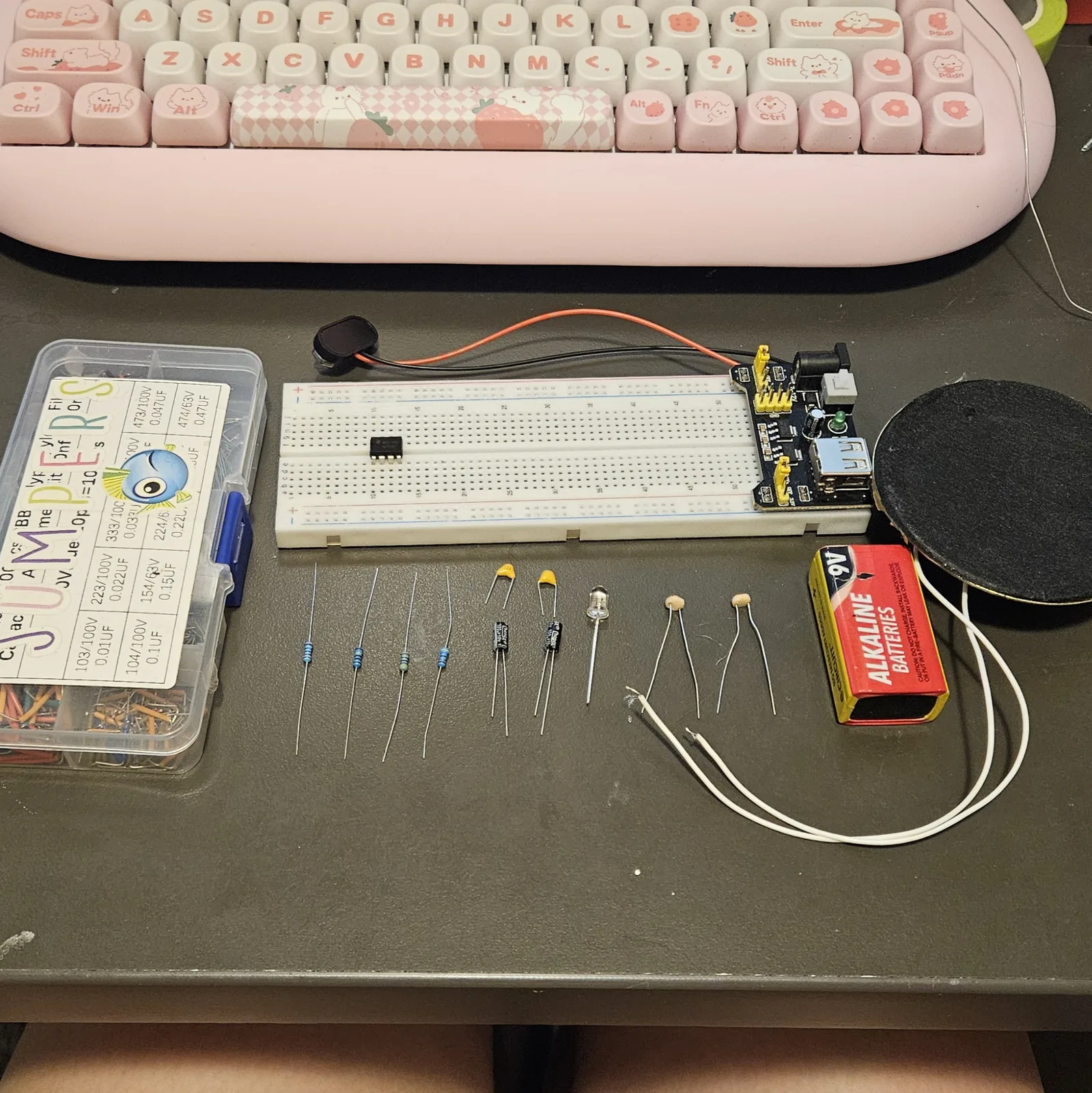 The black 8-pin timer chip seated alone on the white breadboard, straddling the center channel a fifth of the way along, with the rest of the board empty. The remaining parts still lie in a row on the bench below, beside the parts box, the 9-volt battery and the round speaker.