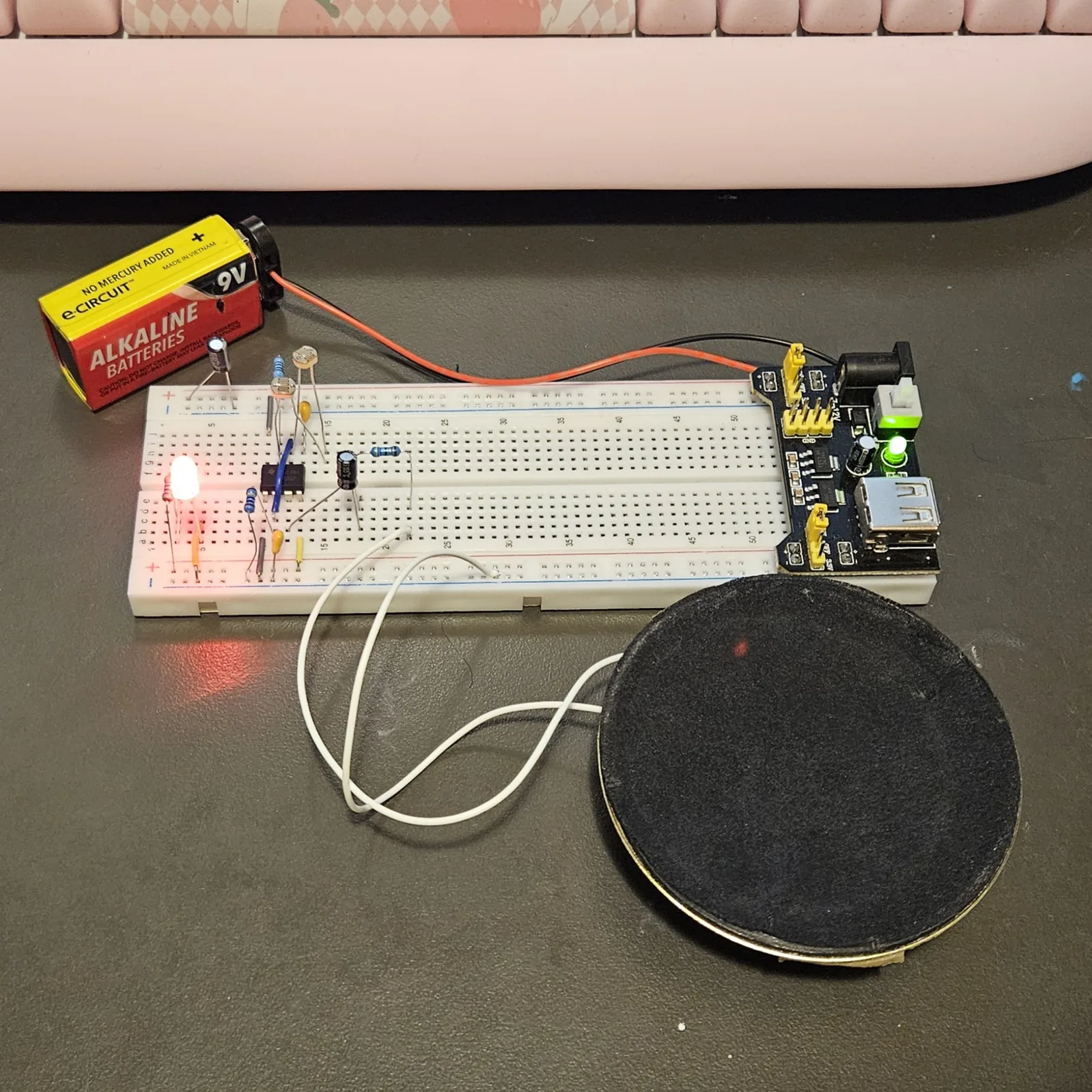 The finished build powered up on a white breadboard on a dark bench. The black 8-pin timer sits across the center channel with a blue jumper arcing over it, a black electrolytic can beside it, yellow ceramic capacitors and banded resistors along the lower bank, and a photocell standing on bent legs at the left. A red LED at the far left is lit and throws a red pool of light across the bench. Two white leads run from the board to a large round speaker lying in front. A black power module is plugged into the right-hand end, fed by a 9-volt battery.