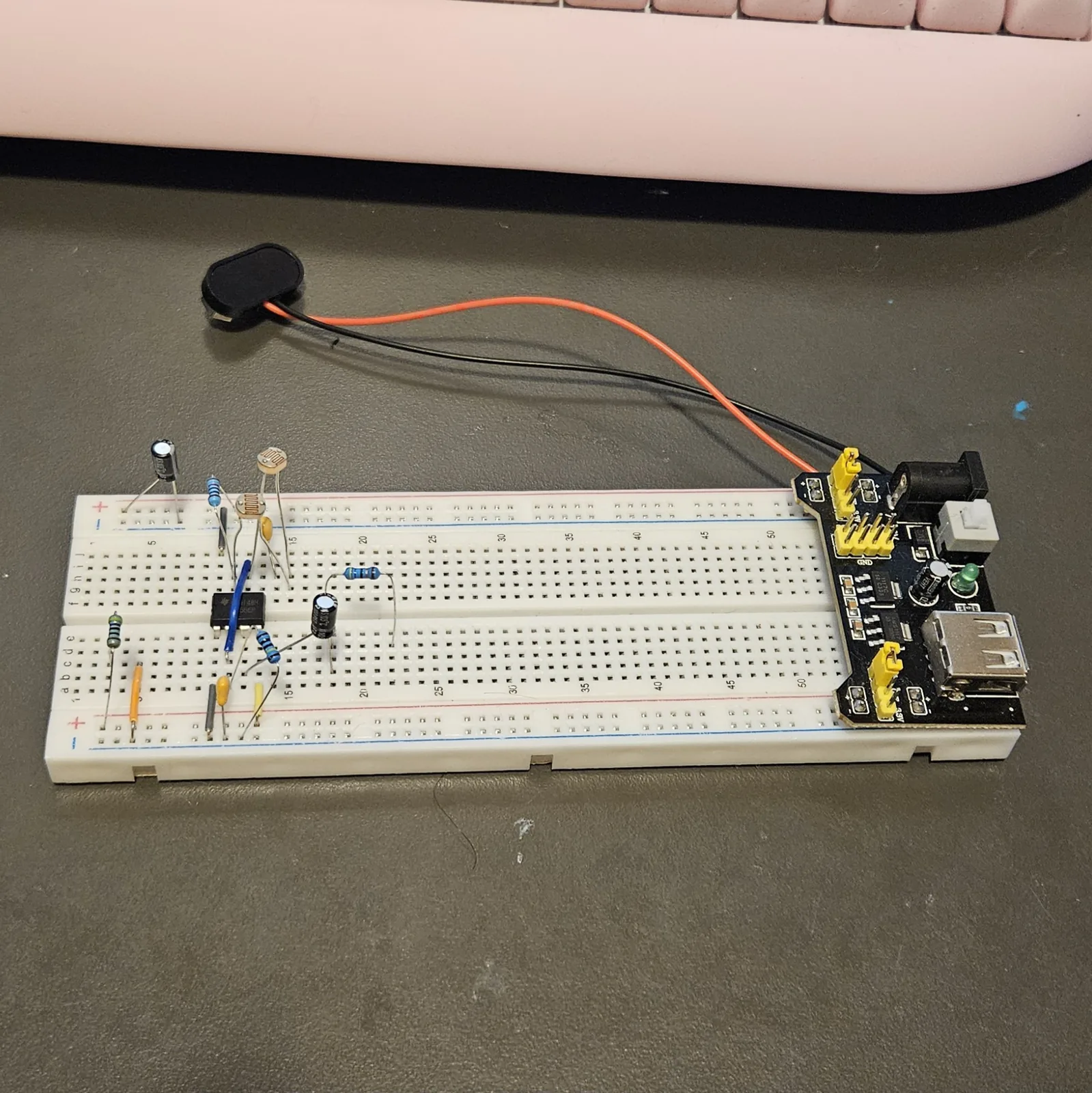 The breadboard with the resistors and a photocell added. A round gold photocell with a wavy trace across its face stands in the upper bank near the chip, with blue-bodied banded resistors either side of it and a green-banded resistor further left in the lower bank. The blue jumper arcs over the black timer chip, and a black electrolytic can and yellow ceramic capacitor sit to the right.