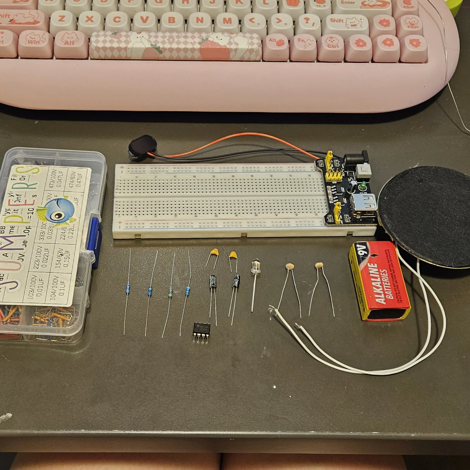 Everything for the build laid out on a dark bench: a compartment box of components at the left, a white breadboard with a black power module plugged into its right-hand end, a red and yellow 9-volt battery, and a large round speaker with a black cone. In front of the board, left to right, sit banded resistors, the black 8-pin timer chip, two round photocells, small yellow ceramic capacitors, two black electrolytic cans and a clear LED, with the speaker's two white leads curling across the bench.