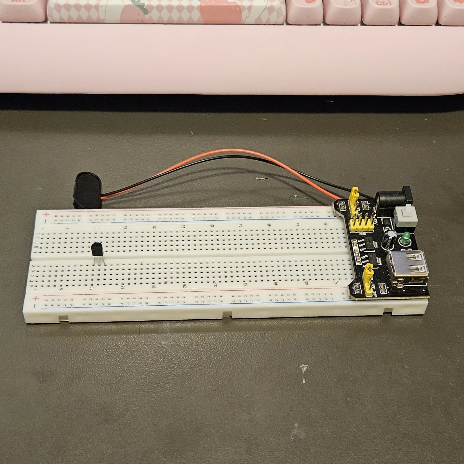 The small black TO-92 transistor seated alone on the empty white breadboard, its three legs pushed into three adjacent rows and its flat face turned toward the camera. The power module and its red and black leads sit at the right-hand end.