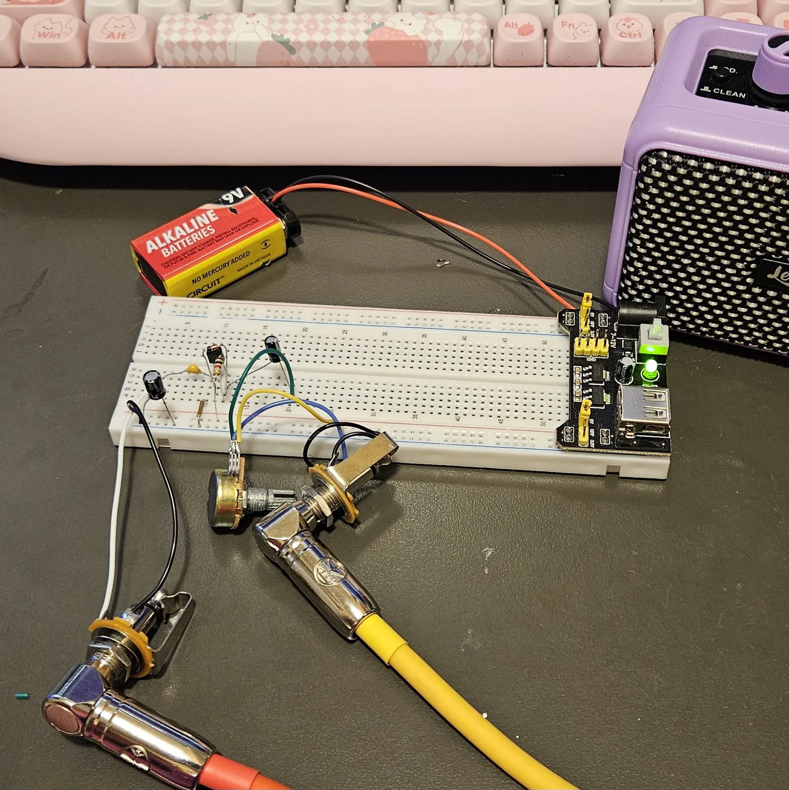 The finished fuzz on a white breadboard on a dark bench, plugged in and playing. A small black transistor, a yellow film capacitor, a black electrolytic can and banded resistors sit along the left of the board, with blue, yellow and green flying leads running down to a potentiometer and two metal jack sockets lying in front. A yellow guitar cable is plugged into one jack and runs to a lilac practice amplifier at the right. A 9-volt battery feeds the black power module at the board's right-hand end, its green LED lit.