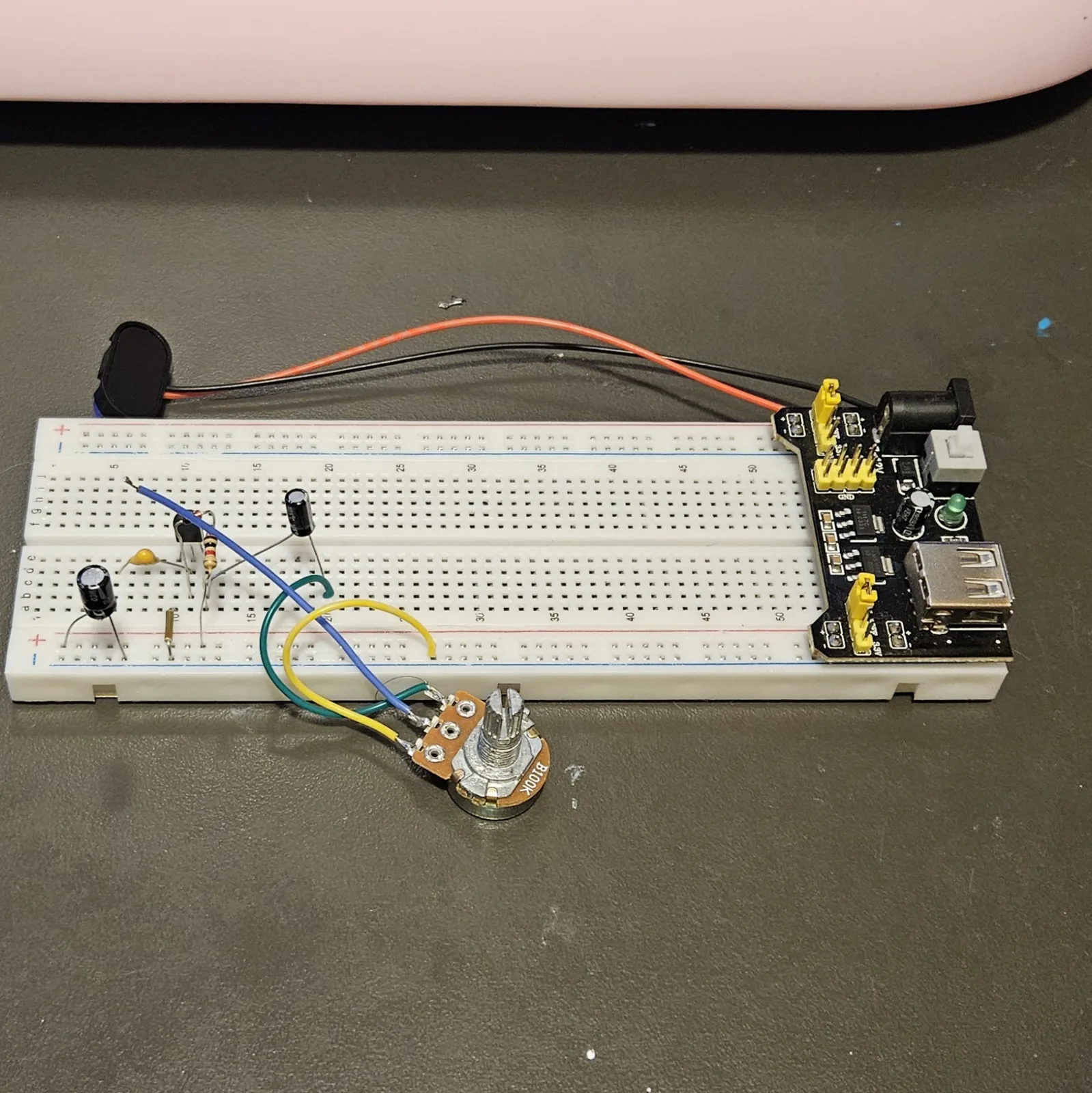The potentiometer now connected to the board by three flying leads in blue, yellow and green, lying on the bench just in front of the breadboard with its shaft pointing up.