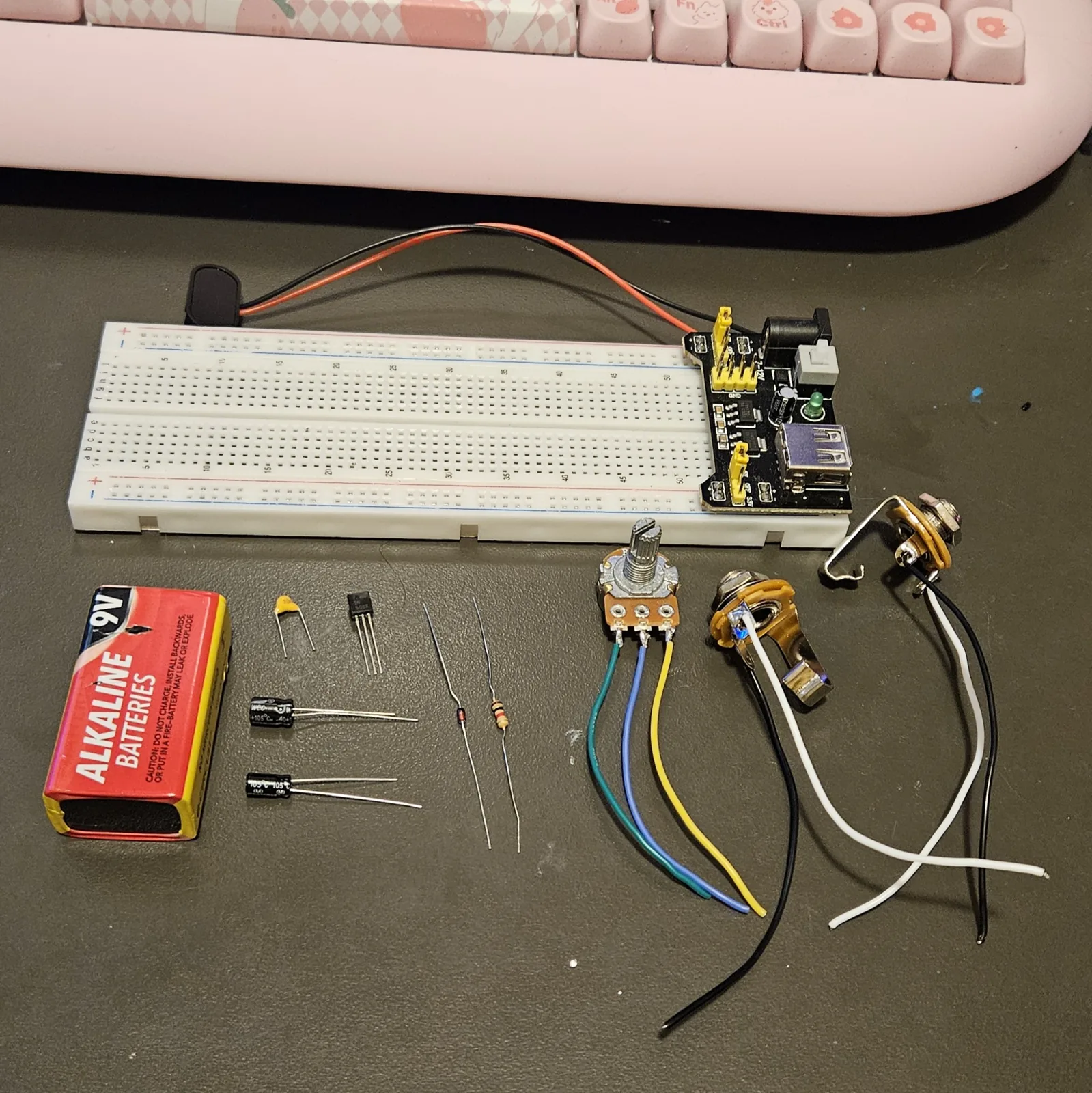 Everything for the build laid out on a dark bench below the breadboard and its power module: a 9-volt battery, a small yellow film capacitor, two black TO-92 transistors, a black electrolytic can, a banded resistor, a glass diode, a potentiometer with blue, yellow and green wires already soldered to its lugs, and two metal jack sockets on white and black leads.