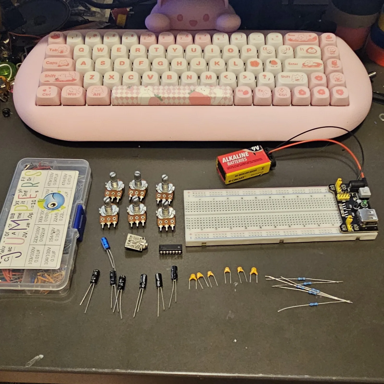 Everything for the build laid out on a dark workbench below a pink mechanical keyboard with strawberry and cat keycaps. On the left, a clear parts box with a hand-lettered capacitor code chart taped to the lid. In the middle, six orange-bodied potentiometers in two rows of three, an output jack, and the black 14-pin CD40106. On the right, a white breadboard with a black power-supply module plugged into one end and a 9-volt battery wired to it. Along the bottom, six black electrolytic capacitors, six small yellow ceramic capacitors, and a row of blue-banded resistors.