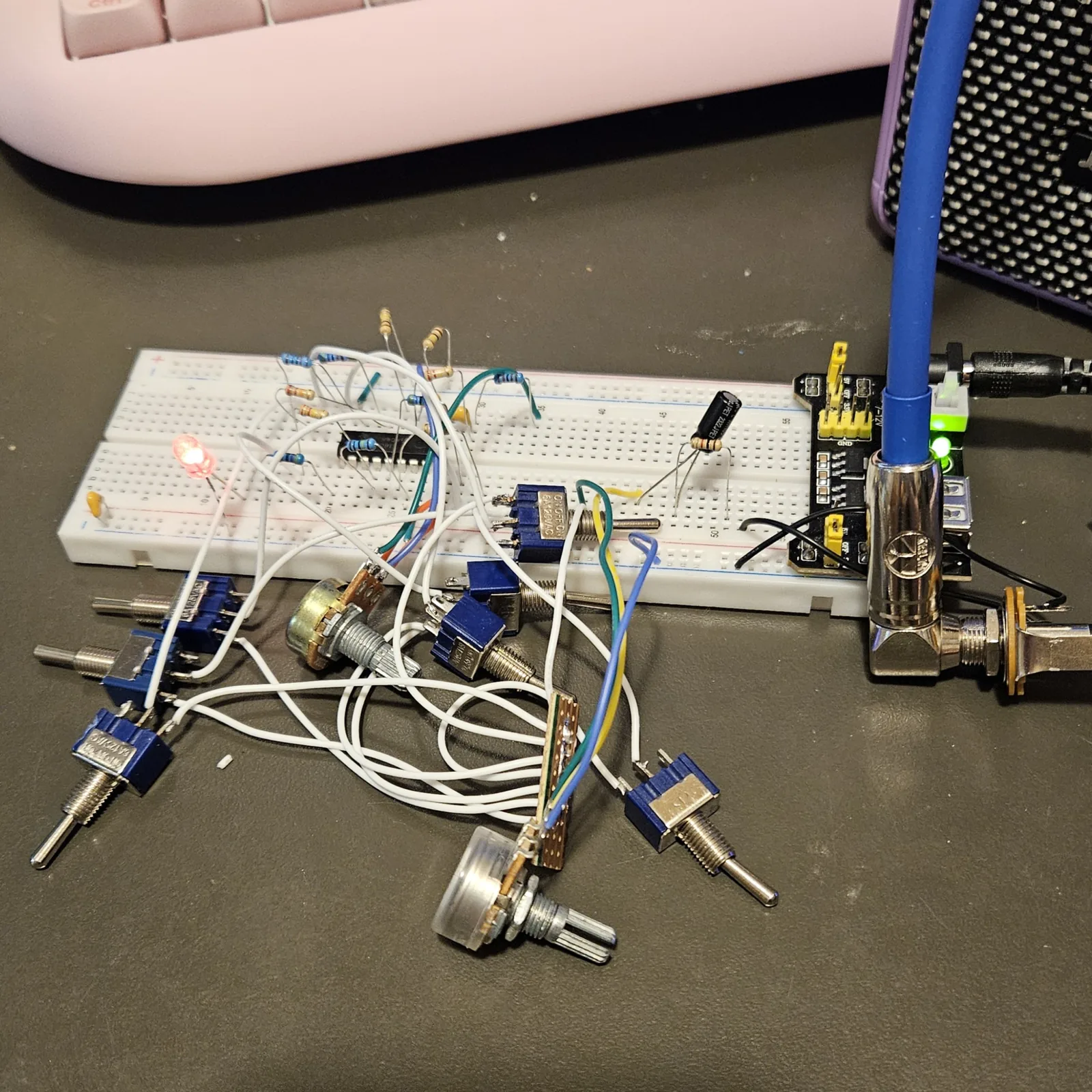 The Master Divider running on the bench. A white full-size solderless breadboard carries the 16-pin CD4060 left of center, with banded resistors standing and arching around it, a small yellow ceramic capacitor at the left end beside a lit LED, and an electrolytic capacitor standing toward the right. A black power module is plugged into the right end of the breadboard with its indicator lit and a barrel plug in it. White, green, blue and yellow flying leads run off the board to a scatter of blue mini toggle switches and two potentiometers lying on the bench in front of it. A blue cable with a chrome right-angle quarter-inch plug runs into a panel jack at the right, and the corner of a speaker grille and a pink keyboard sit behind.