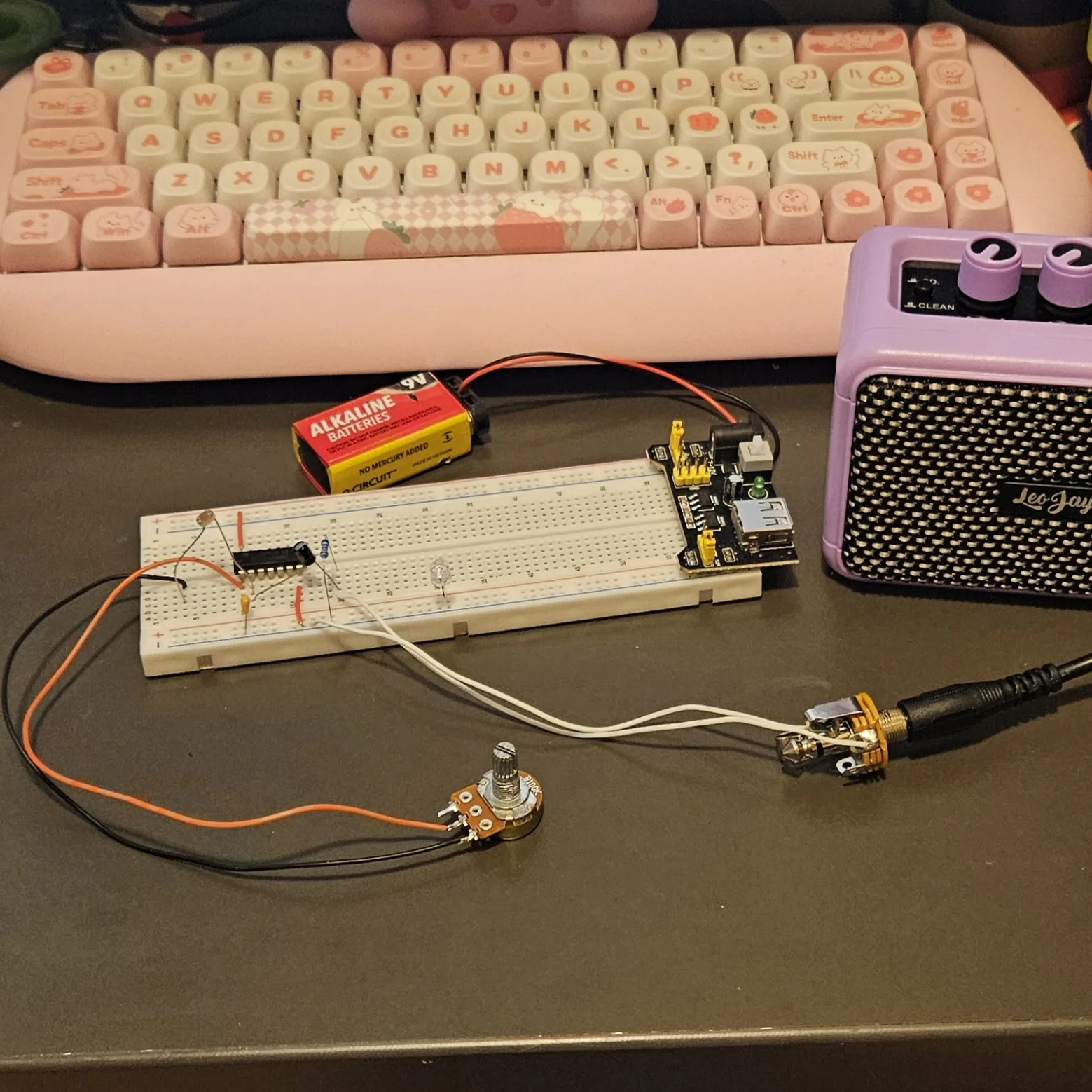 A single-voice optical theremin on a white breadboard on a dark bench. The CD40106 sits across the center channel. To its left stands one round gold photocell with a squiggled face, its legs bent into the board; to its right a single blue-banded resistor, and below the chip one small yellow ceramic capacitor. Two short red jumpers run to the power rails and a clear LED stands further along the board. A 9-volt battery feeds a black power module plugged into the right-hand end. Orange and black wires run off to the left to a potentiometer lying loose on the bench, and two white wires run off to the right to a metal output jack with a black guitar cable leading to a lilac practice amplifier. A pink mechanical keyboard sits behind.
