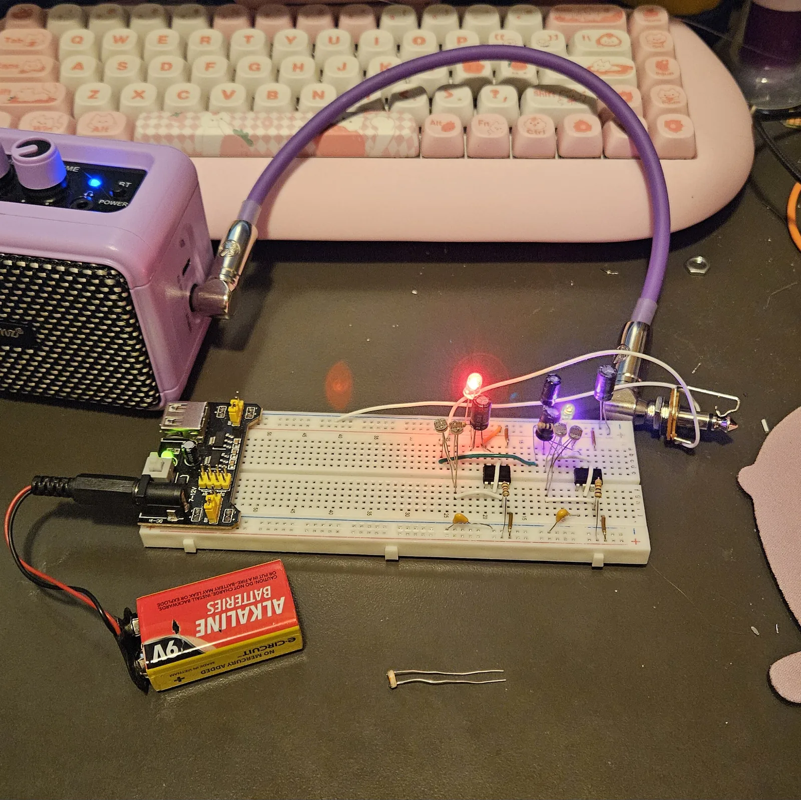 The finished build on a solderless breadboard, running, seen from above on a dark desk mat. A breadboard power supply module sits across the rails at the near end with a black barrel plug in it. Two LEDs on the board are lit, one glowing red-orange and one violet, and between them stand black electrolytic capacitors, banded resistors, two yellow ceramic discs and photocells on bent legs. A red and black lead runs off the near end to a 9V alkaline battery lying on the mat. A purple cable leaves the far end for a lilac practice amp at the top left, which has its own blue power LED lit.
