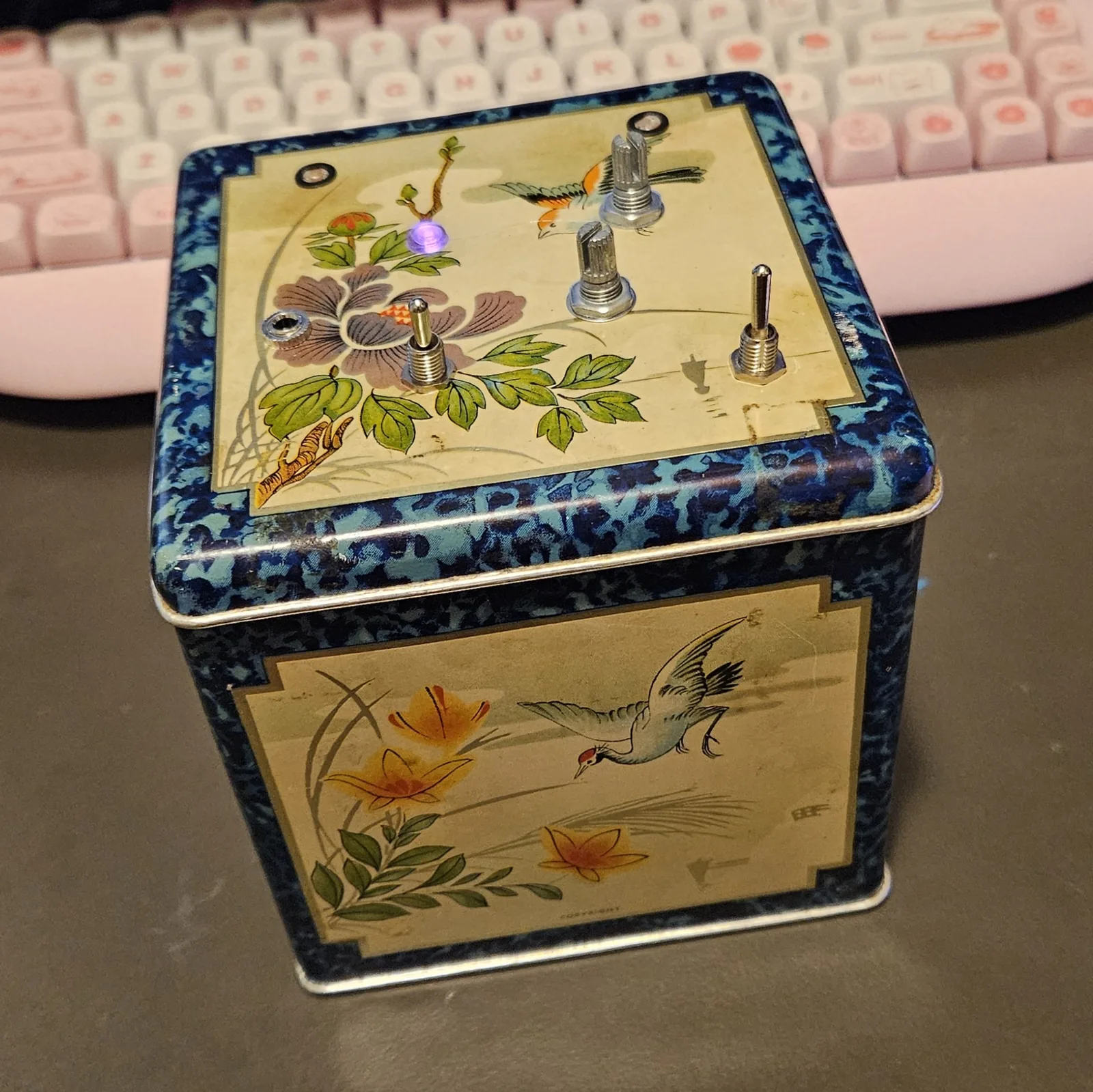 A square tea tin standing closed on a desk, decorated with painted birds and flowers on cream with a mottled dark blue border. Set into the lid are two knurled metal potentiometer shafts with no knobs on them, two small toggle switches, two dark round holes, and a small LED glowing violet.