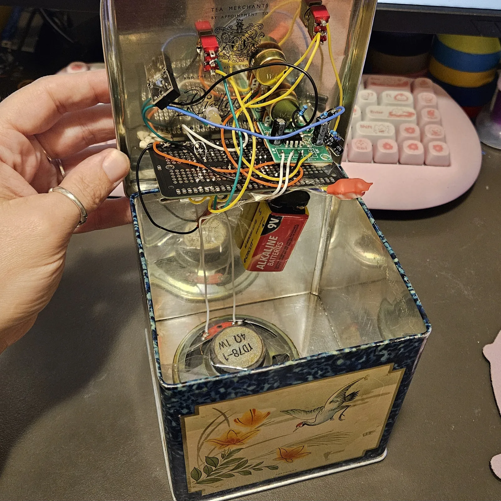 The same tin held open, tipped toward the camera. Under the lid is a dark solderable breadboard crossed by yellow, orange, blue, white and black wires, with a small green amplifier module soldered onto it and the gold cans of two potentiometers standing up behind. A blob of orange putty sits at the rim, a 9V alkaline battery lies in the bottom of the tin, and the tin's printed lining is visible behind the wiring.
