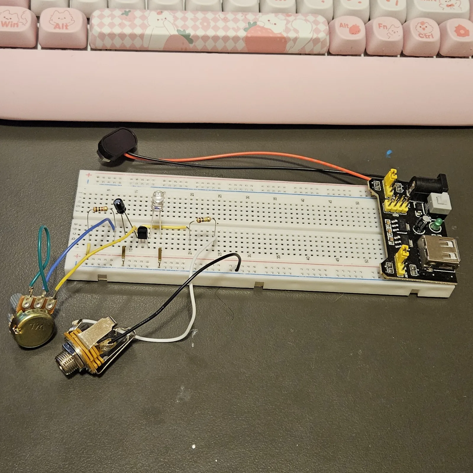 The full build with the off-board parts wired: a potentiometer lying in front of the board with a short green loop soldered between two of its lugs and blue and yellow leads running up into the breadboard, and a metal quarter-inch jack with white and black leads, the white running into the output column and the black into the ground rail. The 9-volt battery snap and its barrel plug rest above the board, not yet connected.