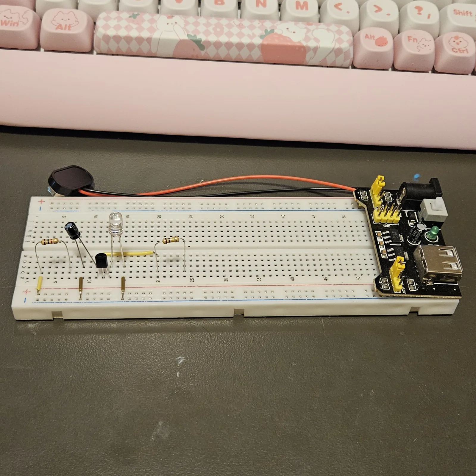 The black electrolytic capacitor added beside the transistor, standing on splayed legs two columns apart with its striped side toward the grounded column. The 1K resistor, the yellow bridge jumper, the LED and the 100K resistor are all in place around it.