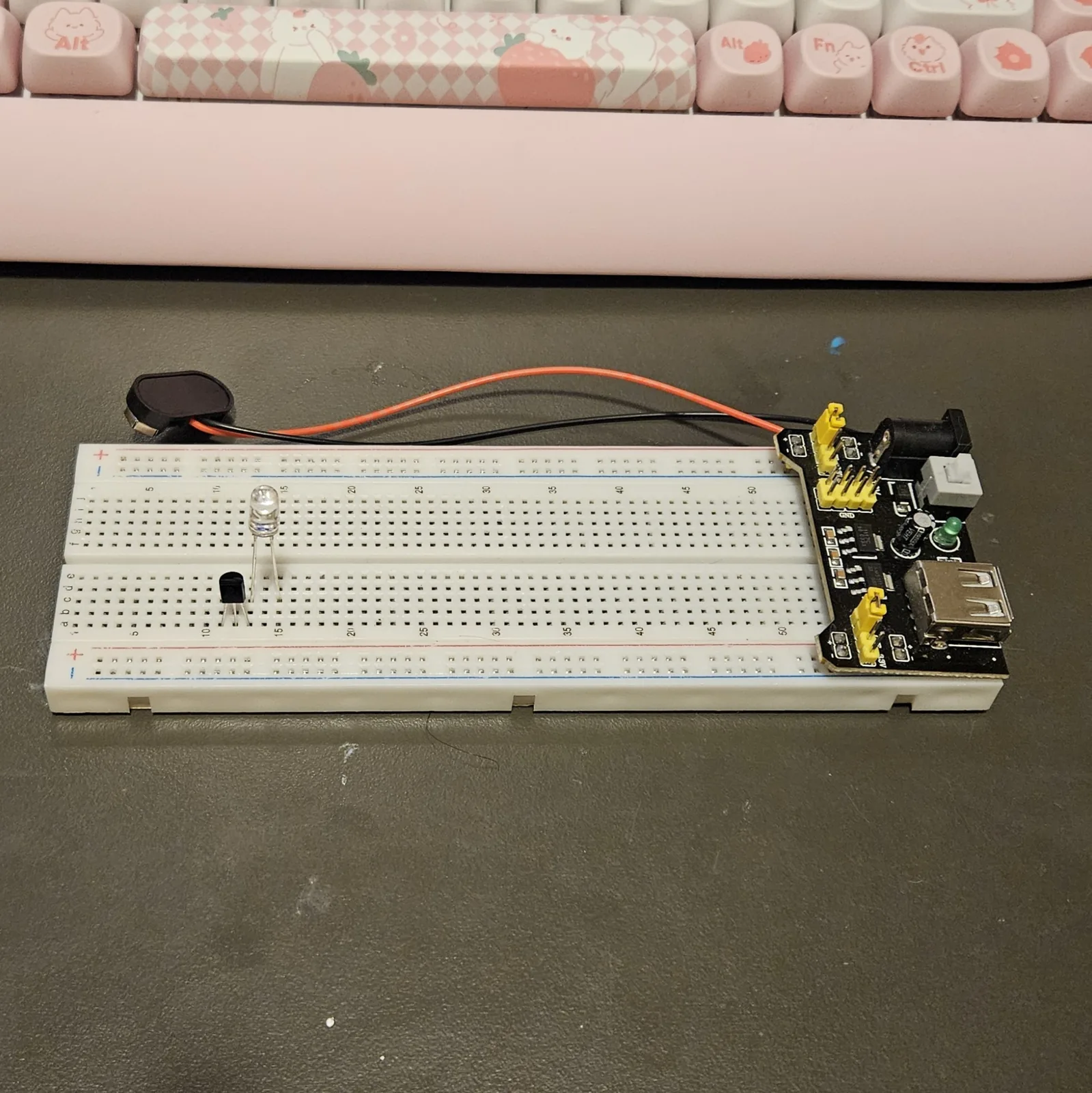 The white breadboard with the first two parts seated in the lower bank: the black TO-92 transistor in three adjacent columns with its flat face toward the camera, and the clear-lensed LED standing two columns to its right. The black power module is plugged into the right-hand end of the board, with its 9-volt battery snap lying unplugged behind it.