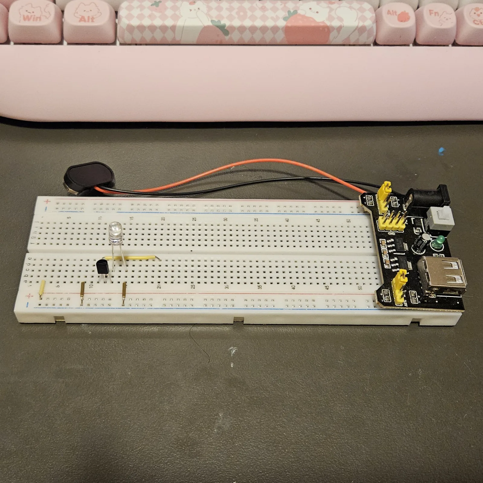 The board with the four jumpers added: three short pins standing in the lower bank and dropping into the bottom rails, one near the left end of the board and two flanking the transistor, plus a yellow jumper lying flat in the row nearest the channel, running from the transistor's columns across to the output area. The transistor and LED stand where they were placed.