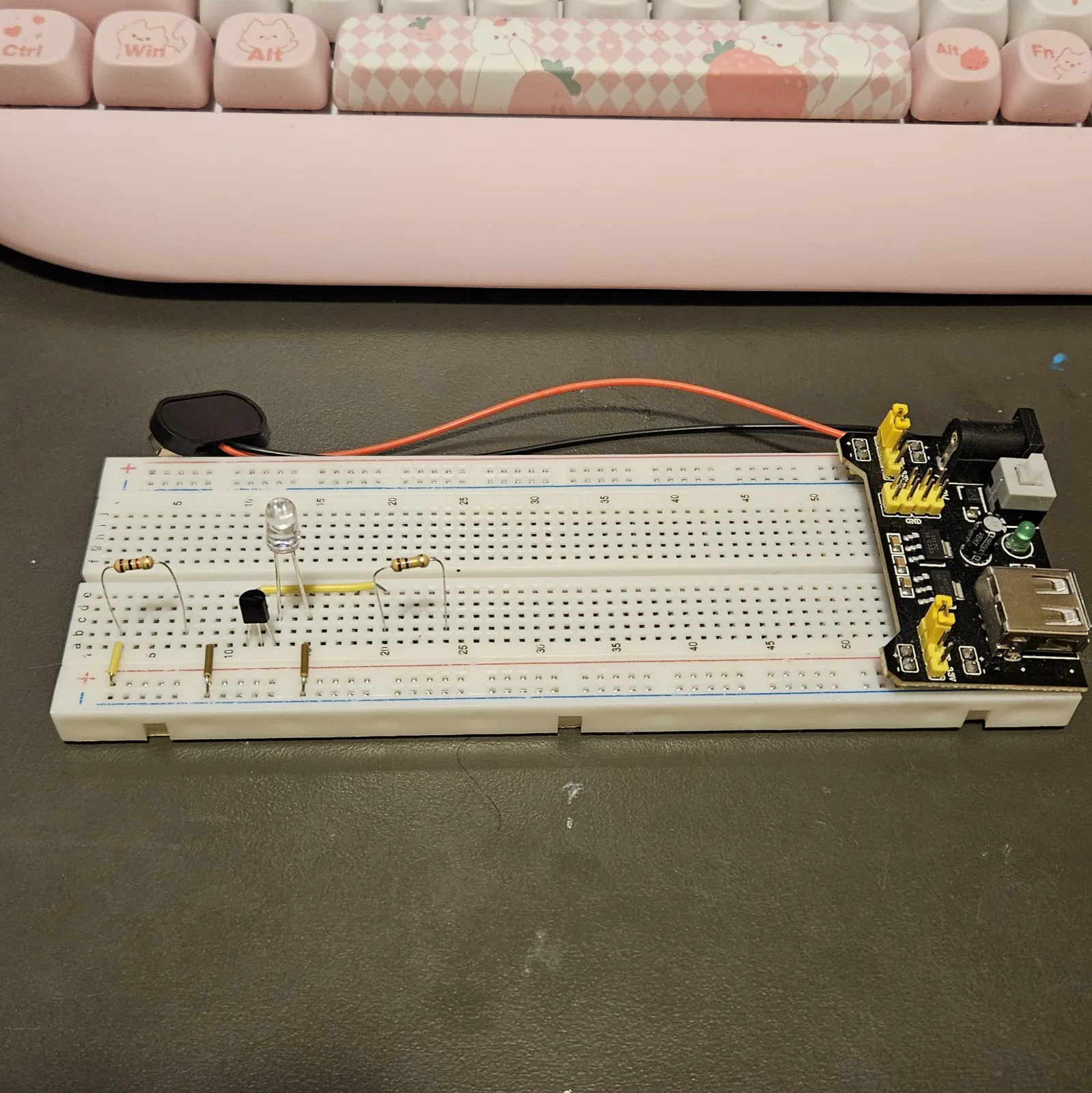 Two banded resistors added to the lower bank: one standing on tall arched legs near the left end of the board on the charging side, and one further right, past the LED, spanning the output columns where the yellow bridge jumper ends. The transistor, LED and jumpers sit between them.