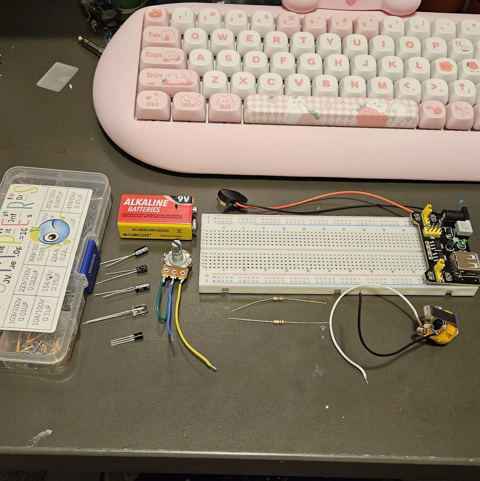 Everything for the build laid out on a dark bench below a pink keyboard: a parts box with a capacitor-code chart on its lid, a 9-volt battery, three black electrolytic capacitors, a clear-lensed LED, a black TO-92 transistor, and a potentiometer with green, blue and yellow wires soldered to its lugs. To the right, a white full-size breadboard with a black power module plugged into its right-hand end, two banded resistors lying in front of it, a 9-volt battery snap with a barrel plug, and a metal output jack on white and black leads.
