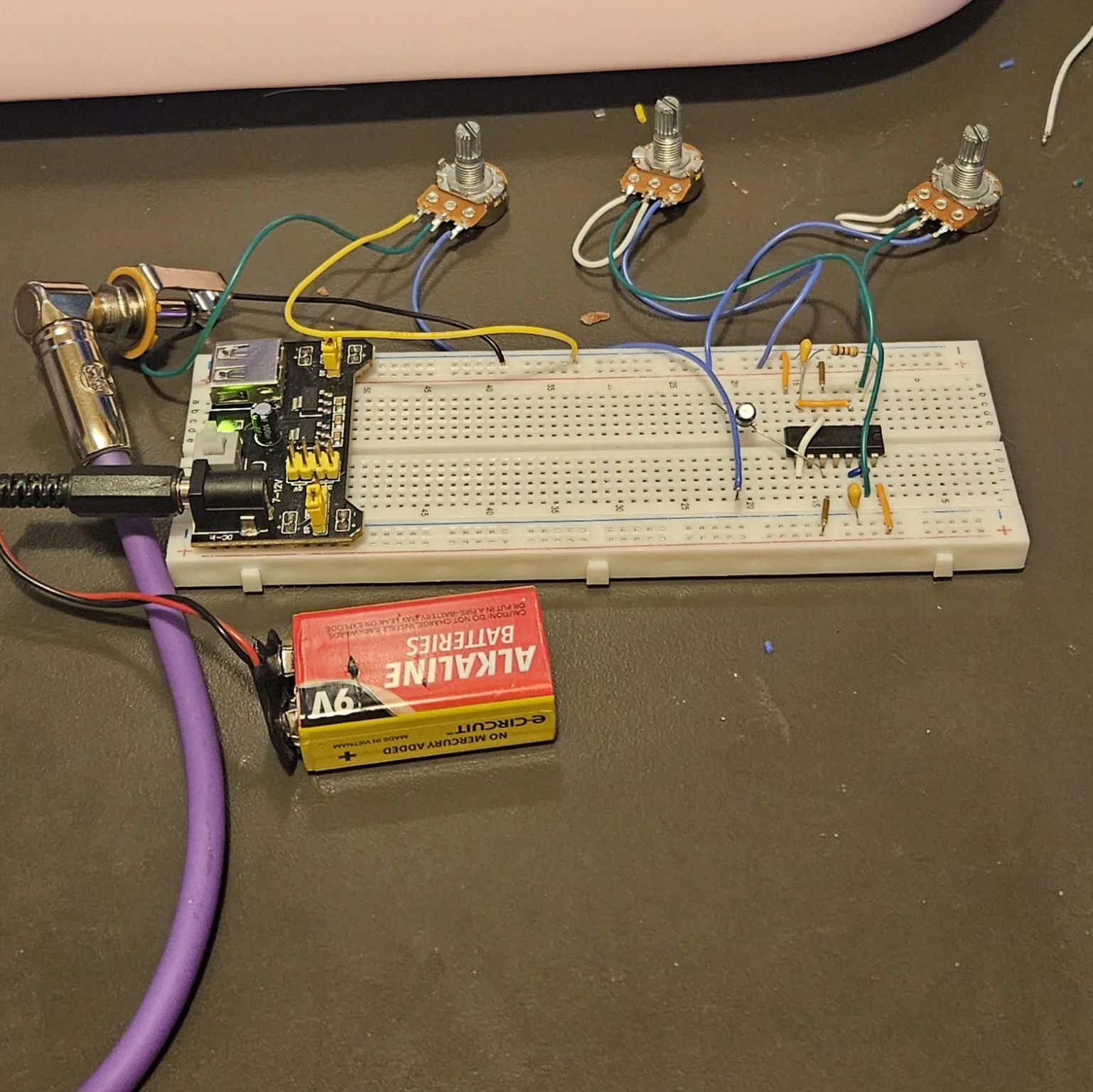 The finished Atari Punk Console running on the bench. A white solderless breadboard carries the NE556 chip near its right-hand end, with banded resistors and small orange ceramic capacitors around it, and blue, green and white flying leads run up to three panel potentiometers lying on the bench above the board. A black power module is plugged into the left end of the breadboard with its green indicator lit, a 9-volt alkaline battery on a snap connector lies below it feeding that module, and a purple cable runs off to the left to a right-angle quarter-inch jack plug.