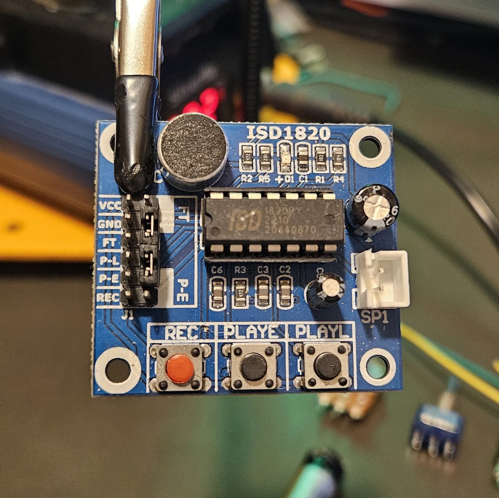 Close-up of the ISD1820 module front. The header along the left edge is silkscreened top to bottom VCC, GND, FT, P-L, P-E, REC. The ISD chip sits in the center marked ISD 1820PY, with the round electret microphone above it, a white two-pin connector marked SP1 on the right for the speaker, and three tactile buttons across the bottom labeled REC, PLAYE and PLAYL.