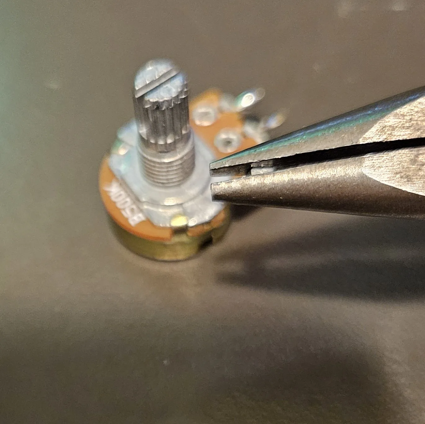 Needle-nose pliers gripping the small anti-rotation tab on the face of the potentiometer to snap it off. The body of the pot is printed B500K.