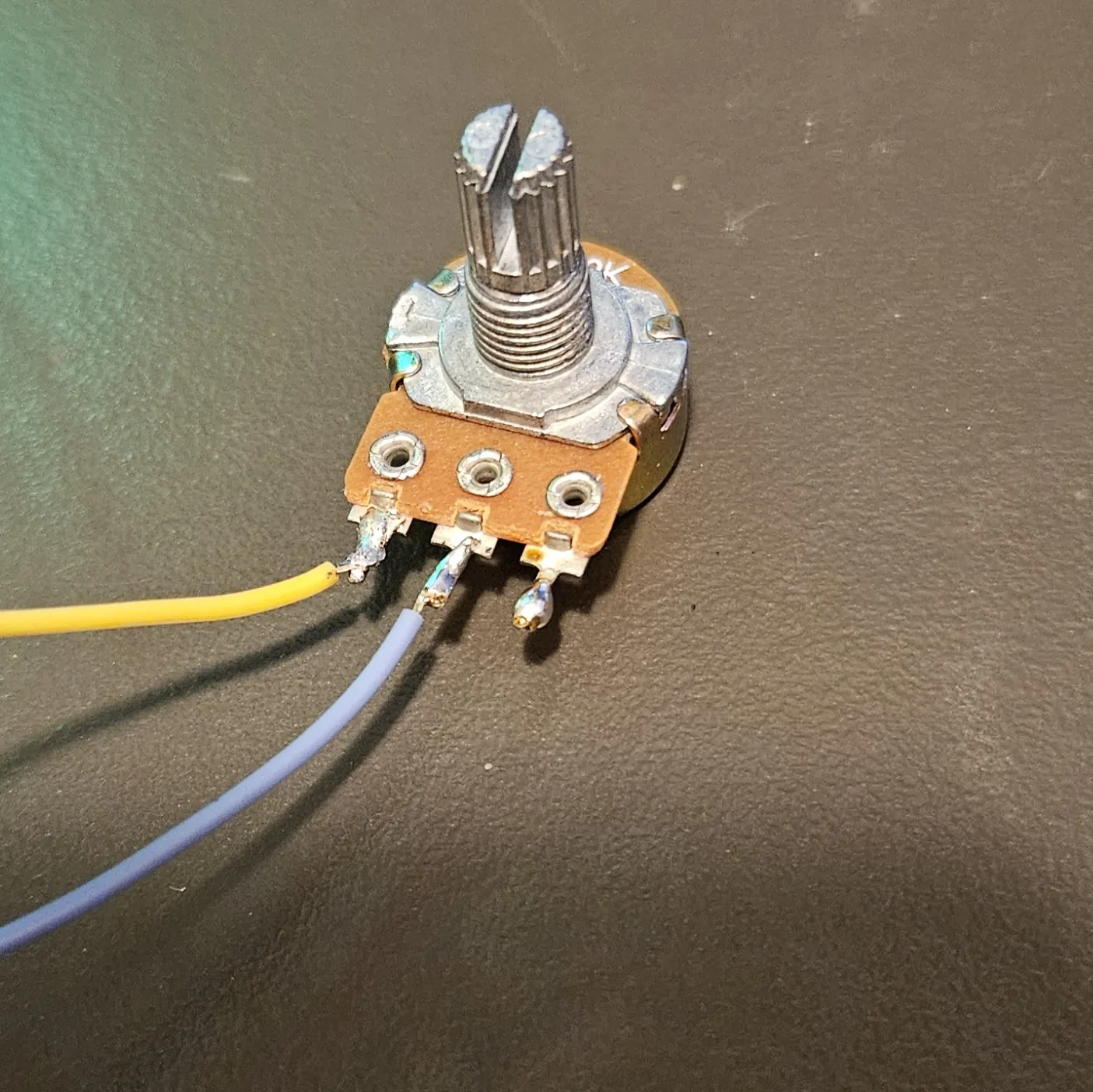 Close-up of the potentiometer with two wires soldered on, a yellow lead on one outer lug and a gray lead on the center wiper lug, the third lug left bare.