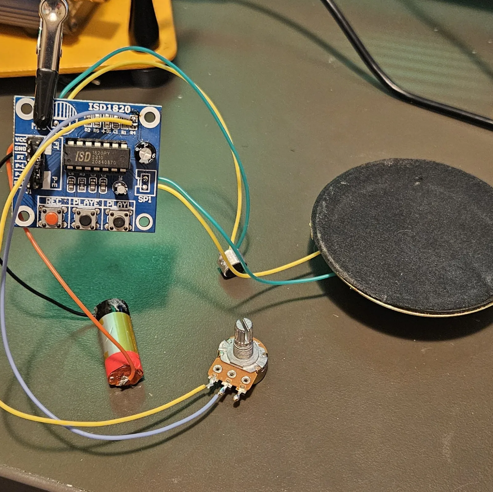 The whole assembly laid out on the bench: the module with its yellow and gray pitch-pot leads running to the potentiometer at the bottom, the electret mic and speaker on green and yellow leads, and the lithium cell alongside.