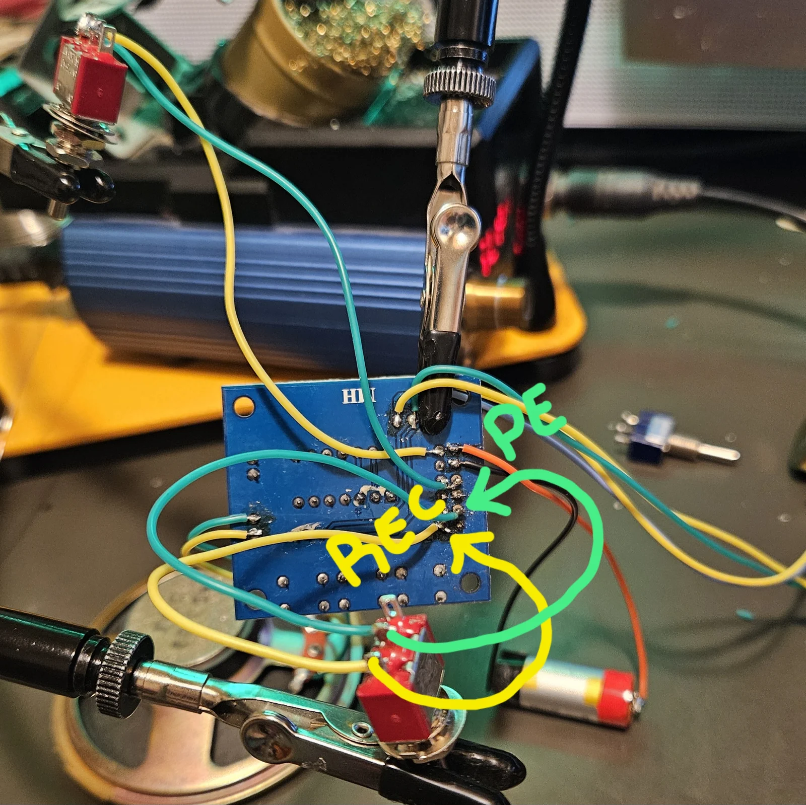 The underside of the ISD1820 module wired for the working build, held in helping hands, with hand-drawn annotations added. A green arrow labeled P-E points to a pad on the right-hand pad row, and the teal wire leaving it is traced in green down to a red toggle switch at the bottom. A yellow arrow labeled REC points to an adjacent pad, and its yellow wire is traced down to the same switch area. Orange and black power leads run off to a cylindrical lithium cell on the right.
