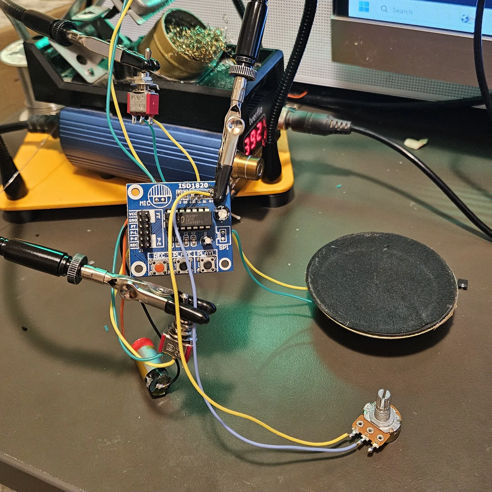 The complete build spread across the workbench: the ISD1820 module clipped in helping hands with its J1 header labels visible, two red toggle switches wired in, the lithium cell, the relocated electret microphone, the large speaker, and the pitch potentiometer, all connected with colored hookup wire.
