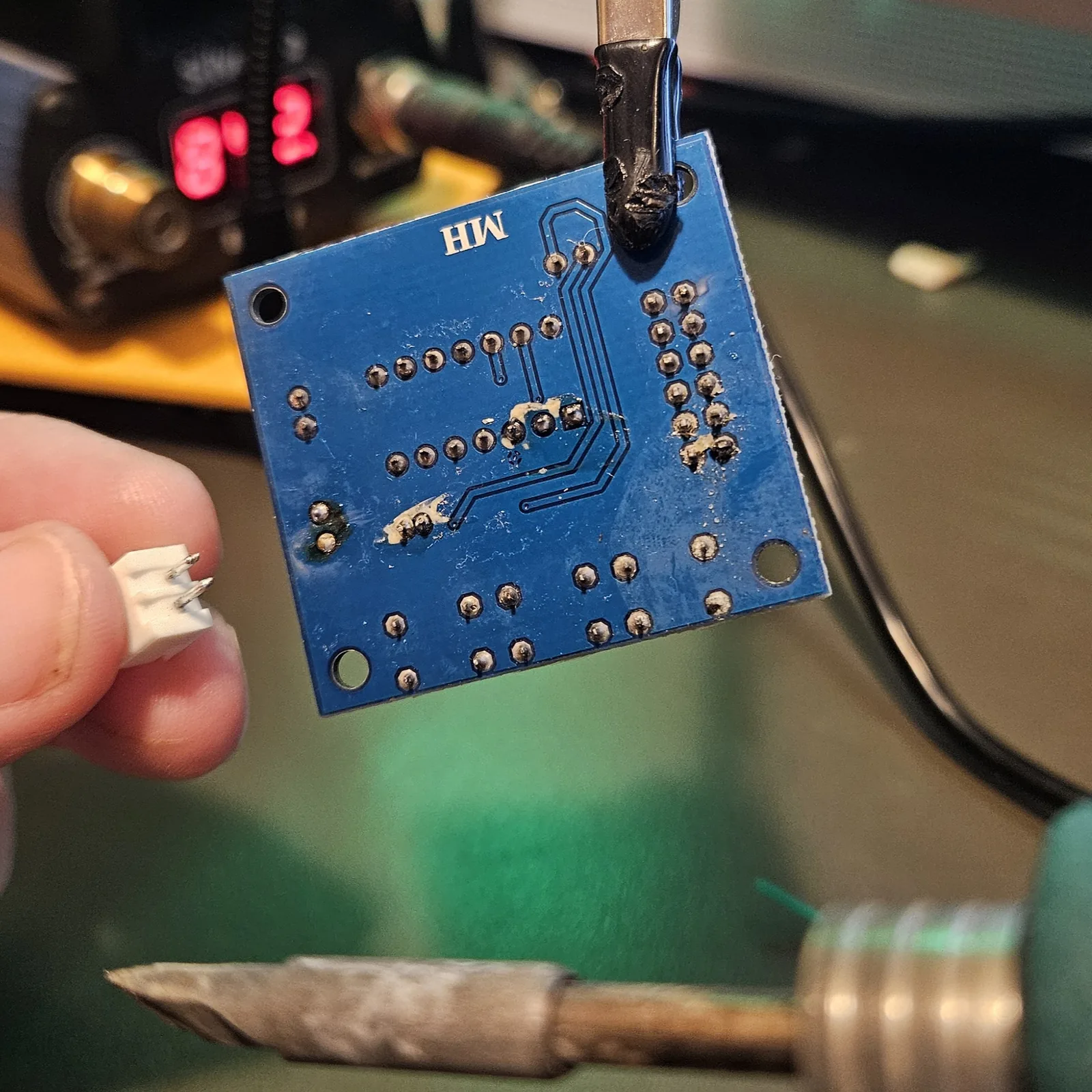 The white two-pin SP1 speaker connector now removed and held between finger and thumb beside the board. The pads it came off are clear, wet-looking with pale flux residue, and undamaged.