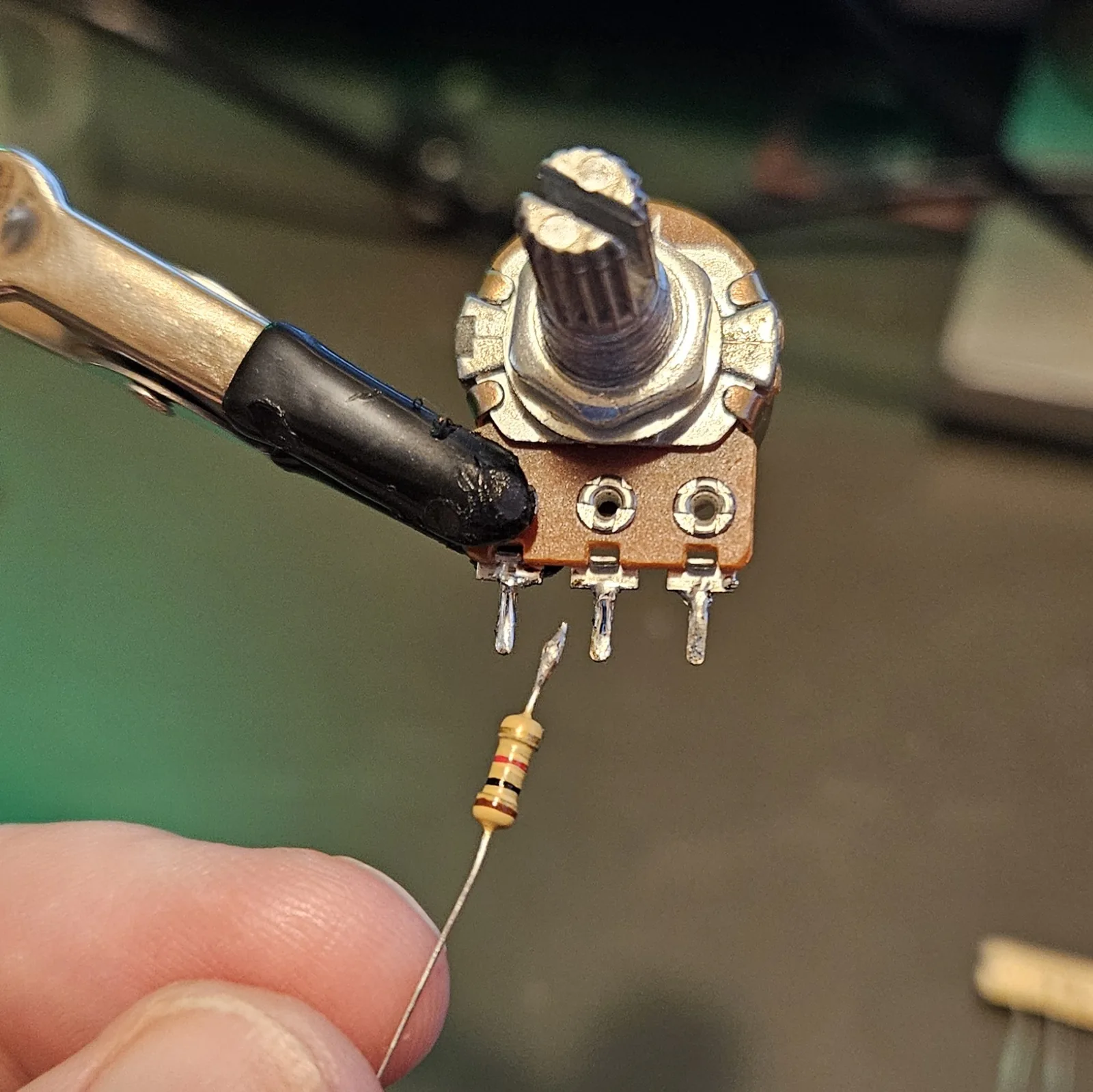 A color-banded resistor held in a clip below a potentiometer lug, its lead freshly tinned. The bands read brown, black, red, gold.