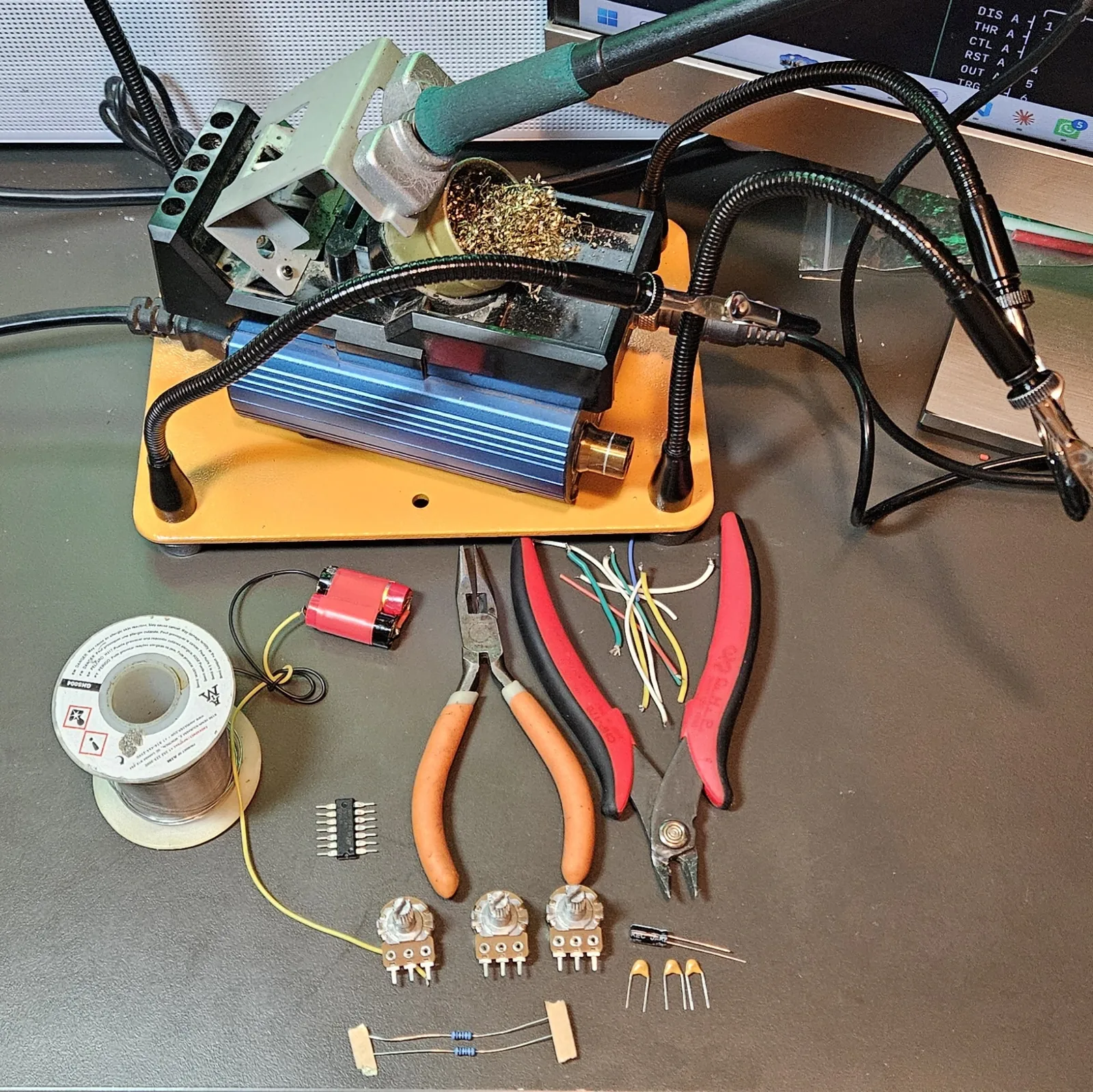 Everything laid out on the bench: a soldering station and helping-hands clamp at the top, a spool of solder, three pairs of pliers (needle-nose, flush cutters, side cutters), a coiled red vape battery pack with yellow and black leads, the NE556 chip, three potentiometers, an electrolytic cap, three yellow ceramic caps, two blue resistors taped together, and a bundle of multicolored hookup wire.