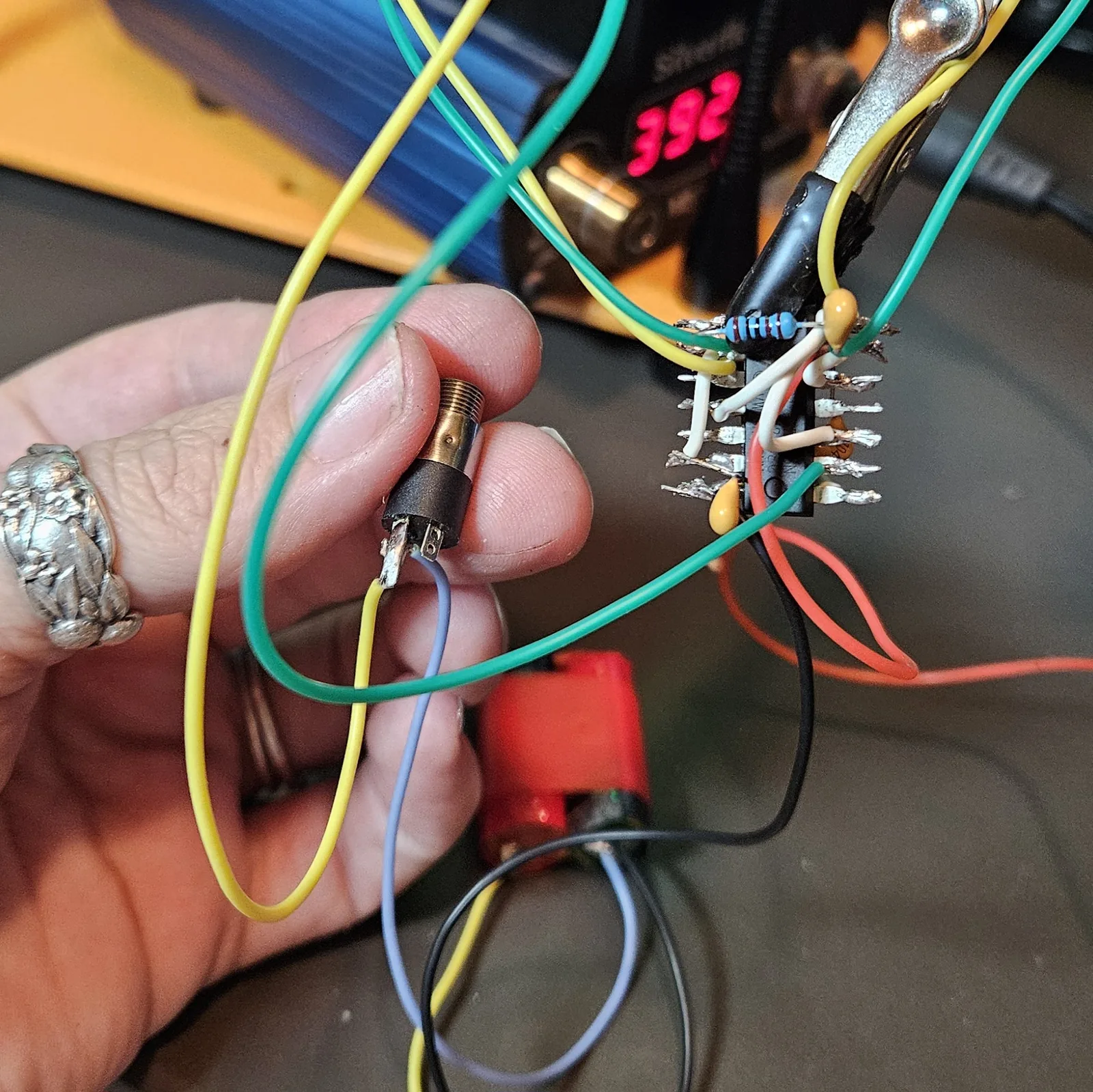 A hand holding a small metal panel-mount output jack wired with two leads, next to the fully wired dead-bug NE556; the soldering-station readout shows 392.
