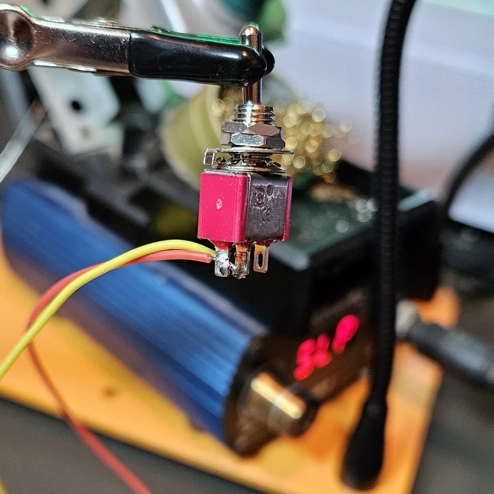 A small red-and-black SPST toggle switch held in the helping-hands clip, with a yellow wire and a red wire soldered to its two lugs; the soldering-station temperature readout glows in the background.