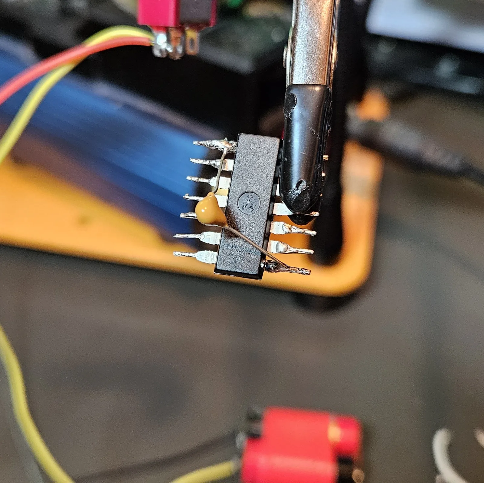 A yellow 0.1 microfarad ceramic decoupling capacitor and a short bare jumper soldered across the NE556's power pins, the iron tip resting on a joint.