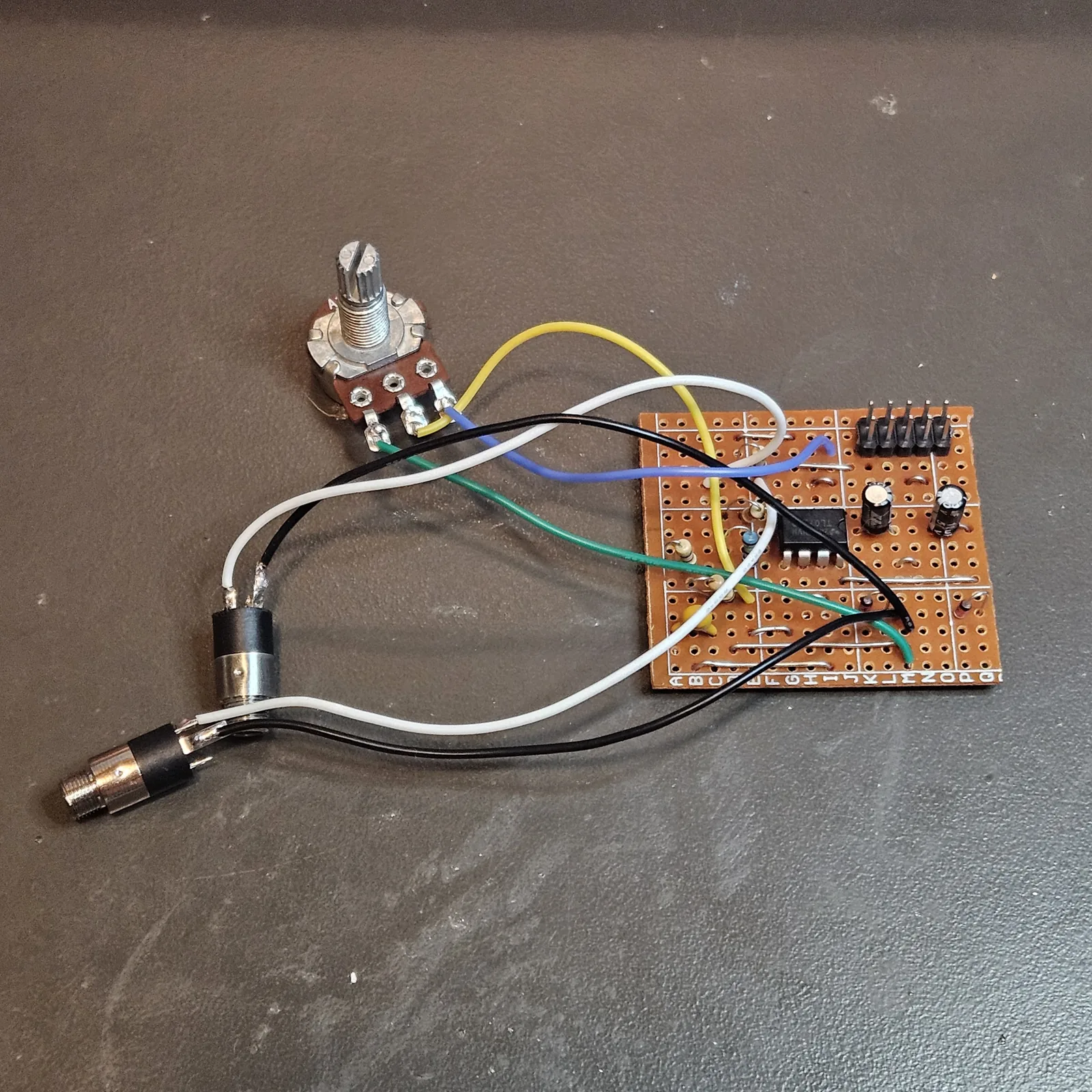 The Output Rack built on a small square of stripboard, lying on a dark bench. The board carries the ten-pin power header at one corner with two small black electrolytic capacitors beside it, an 8-pin chip in the middle, a few resistors, a small yellow capacitor and a row of bare wire links, with the strip letters A to Q printed along one edge. Yellow, blue, green, white and black flying leads run from the board to a volume pot with a knurled shaft and to two small panel jacks lying loose beside it.