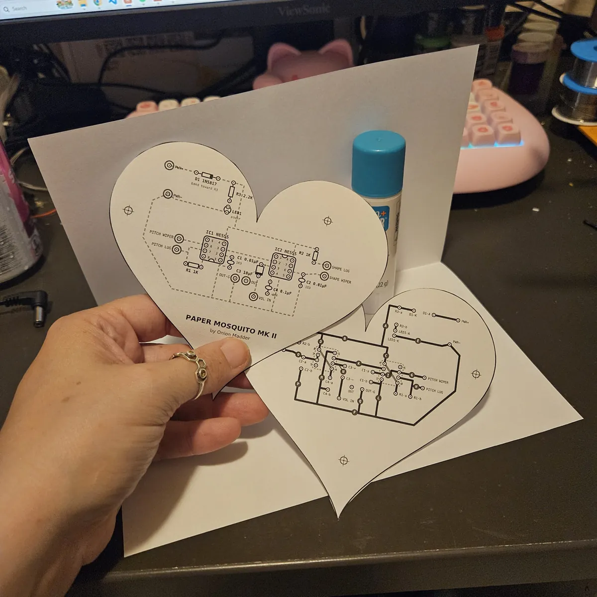 Two paper hearts cut out separately: the FRONT heart, printed with the parts and the title Paper Mosquito Mk II, held up in a hand, and the BACK heart with its black traces lying on a white sheet. A blank sheet of paper stands folded behind them, with a blue glue stick on the dark desk.
