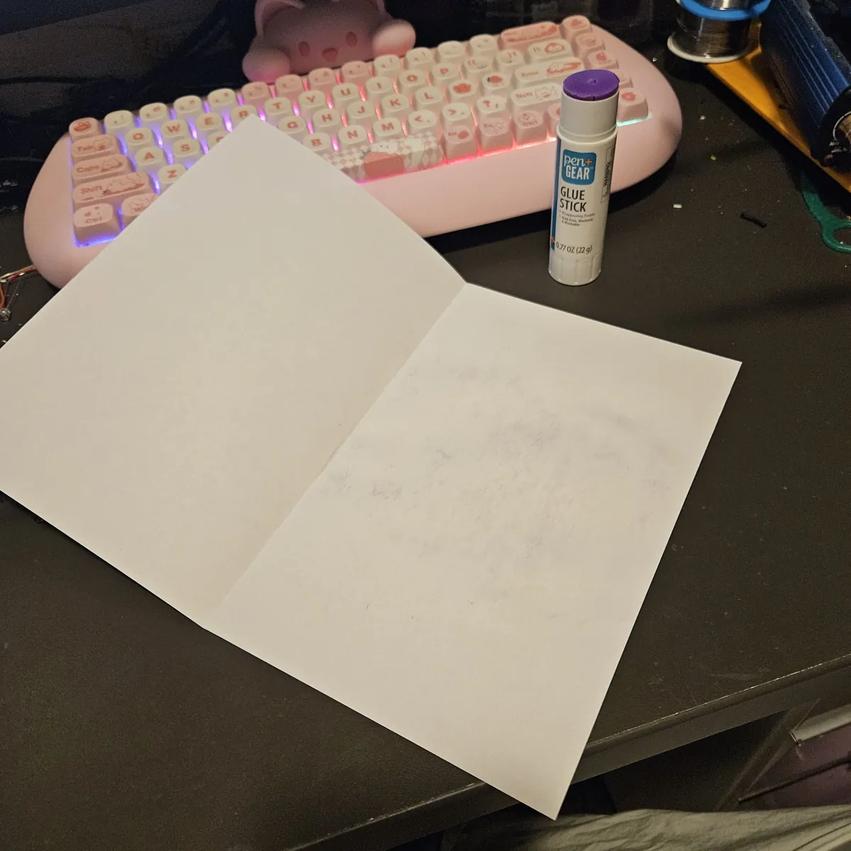 A blank sheet of printer paper folded in half and lying open on a dark desk, glue stick rubbed over one inside half. A glue stick and a pink keyboard sit behind it.