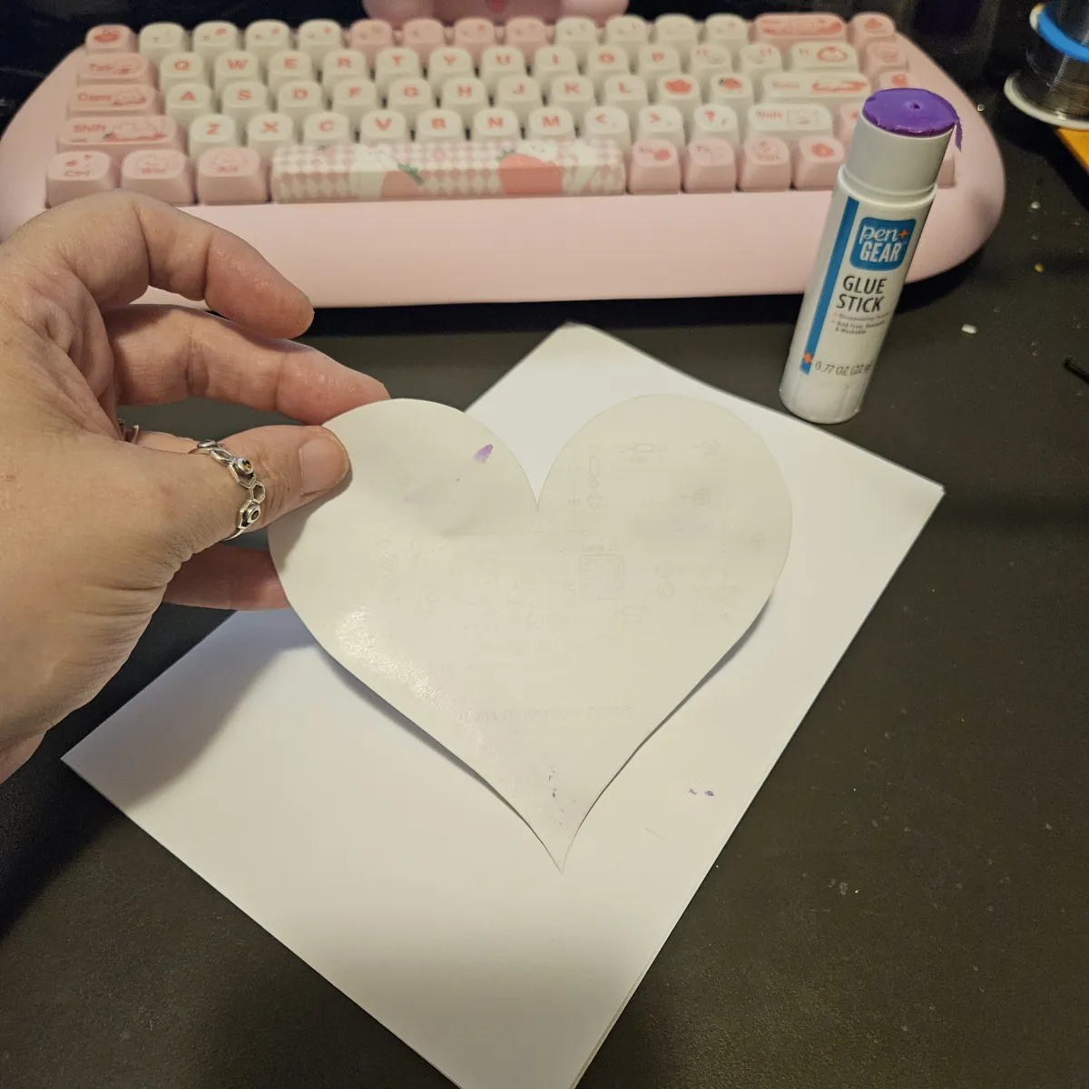 A hand holding a paper heart by its edge, its unprinted back facing the camera and shiny with glue, over the folded core sheet.