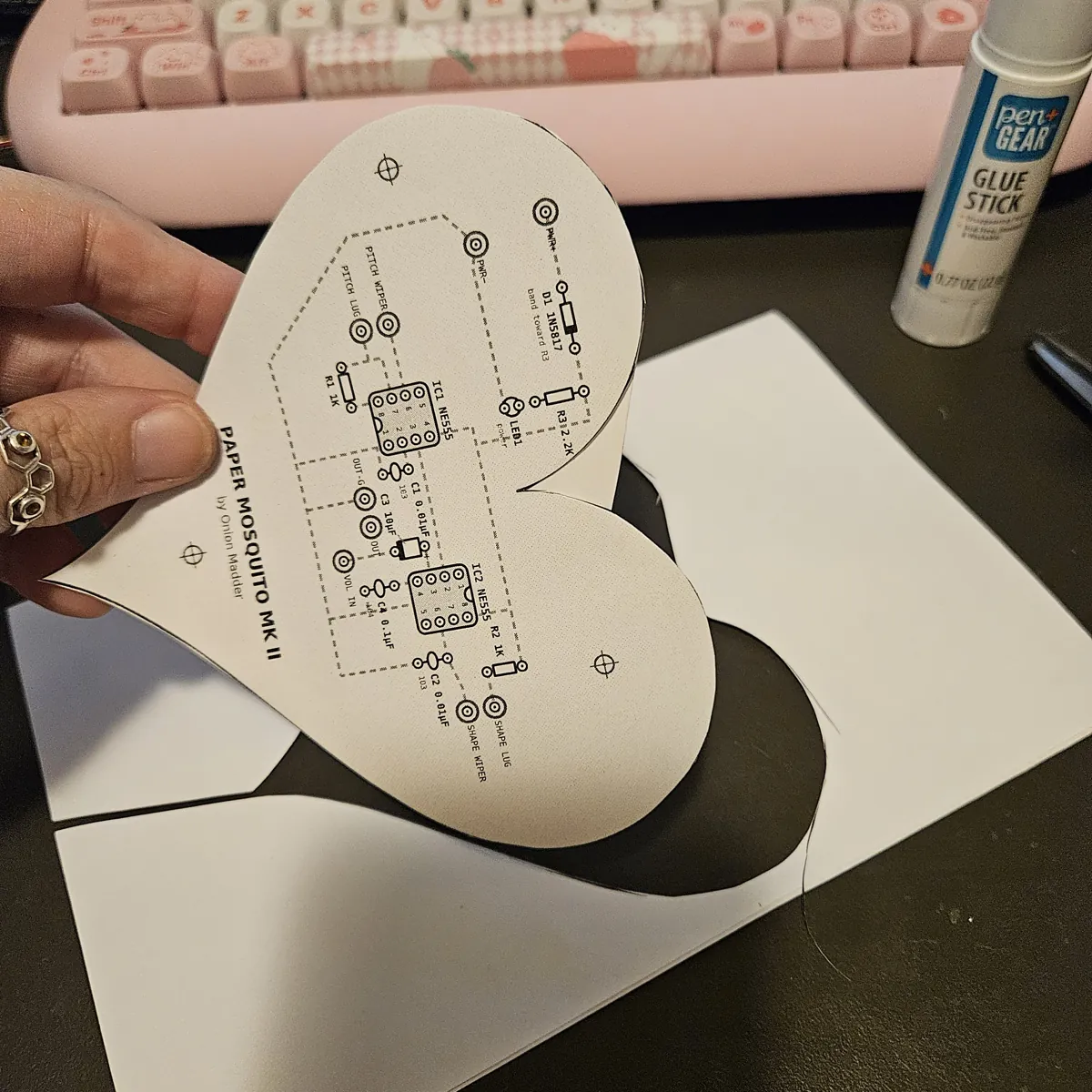 A hand lifting the FRONT heart, now cut out together with the core behind it, from the leftover core sheet, which has a heart-shaped hole in it.