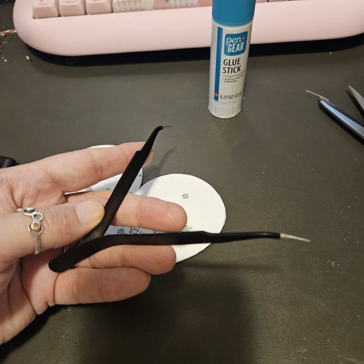 A hand holding the black tweezers pinched closed beside the heart, so the two points meet in one tip.