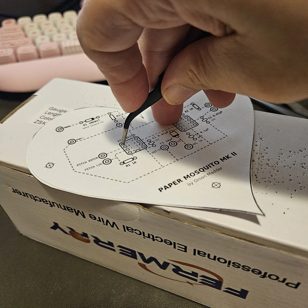 The heart lying front up on a white cardboard box, a hand pushing the closed tweezers down through a chip hole. The box beside the heart is already dotted with little holes from the poking.