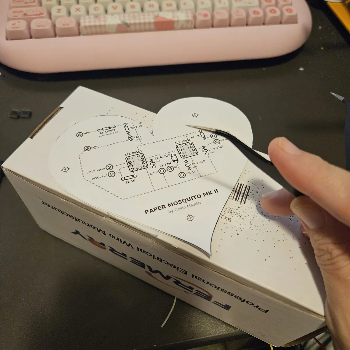 The heart on the cardboard box, a hand poking the holes along the right side with the tweezers.