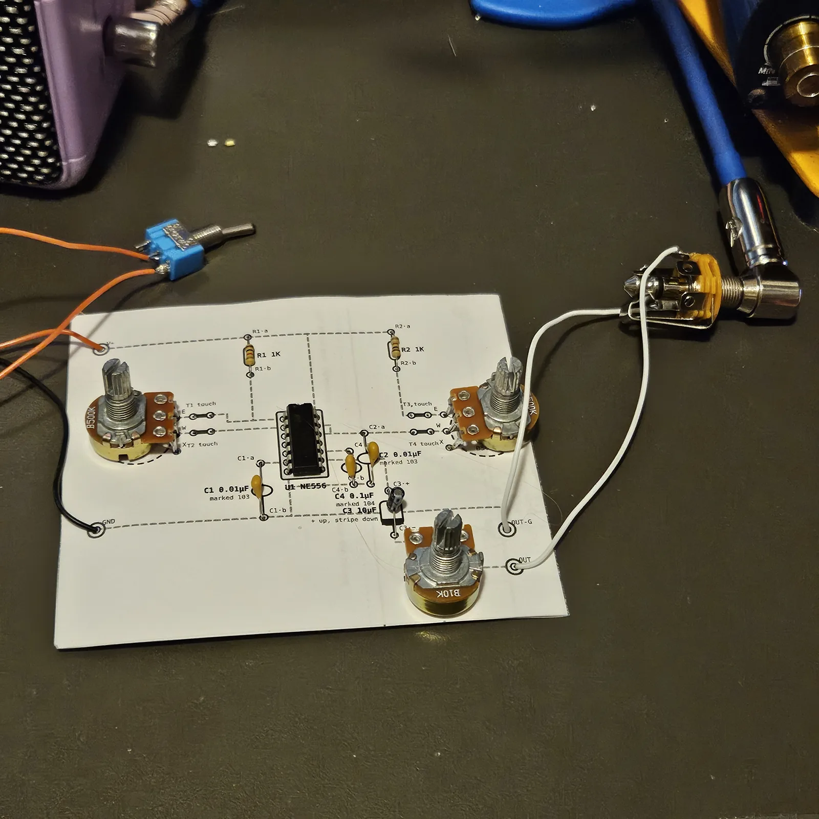 The finished paper circuit on a dark bench mat: a white printed sheet glued over card, with three potentiometers pushed through it, two at the sides and a B10K at the bottom. A black 14-pin chip sits in a socket in the middle, two 1K resistors with brown, black and red bands stand near the top edge, and three small yellow ceramic capacitors and a little black electrolytic sit beside the chip. The part outlines, values and dashed connection lines are printed on the paper around each part. A blue mini toggle switch on orange wires hangs off the top left corner, a black wire runs into the GND hole, and two white wires run from the OUT and OUT-G holes to a quarter-inch jack at the right with a cable plugged into it.