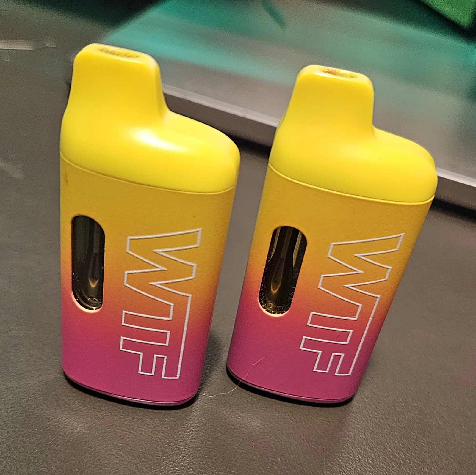 Two yellow-and-pink 'WTF' brand disposable vapes standing side by side on the bench.