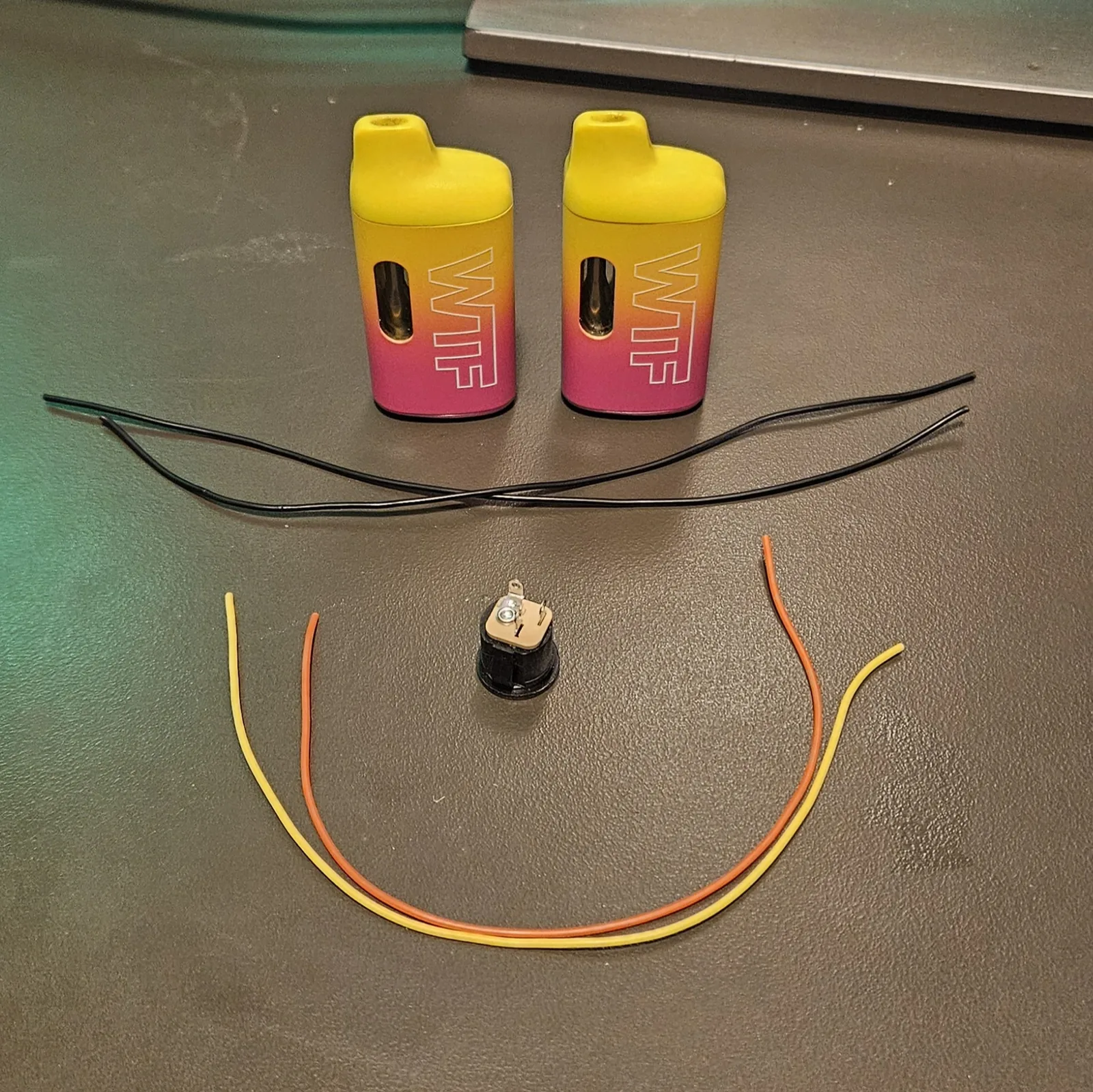 The parts laid out on a cutting mat: two black ground wires up top, an orange and a yellow lead wire curving below, and one small salvaged lithium cell in the middle.