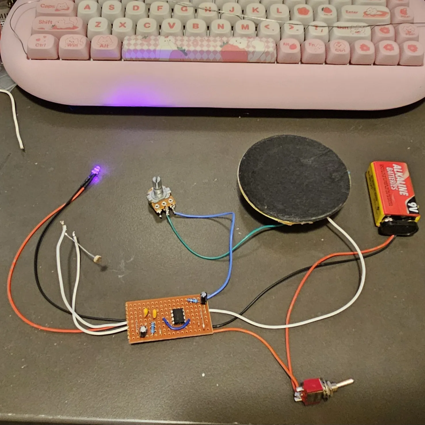 The finished theremin on a dark bench below a pink keyboard: a small copper-colored stripboard carrying the 8-pin timer chip, two yellow ceramic capacitors, two black electrolytic cans, a banded resistor and a blue link wire, with flying leads running out to a large flat speaker, a photocell on white wires, a 9-volt battery on red and black leads, a toggle switch on orange wires, a potentiometer, and a small purple LED, lit.