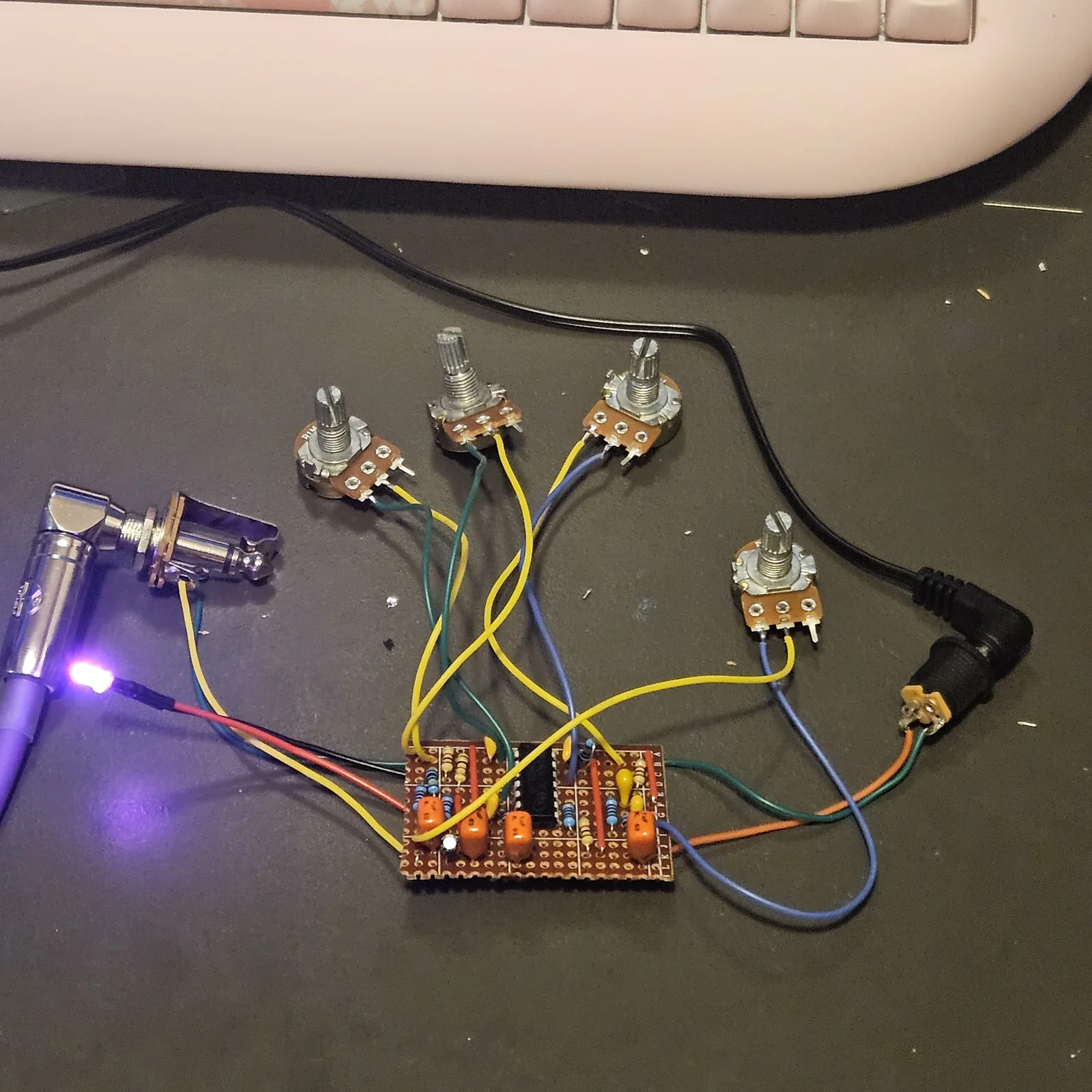 The 12 by 18 board wired up loose on a dark desk mat, not boxed, with a pink keyboard across the top of the frame. The stripboard sits low in the middle, its strips lettered A to L down the right edge. A black 14-pin chip stands in the center, with four orange box capacitors along the lower half, yellow drop-shaped ceramic capacitors, blue and tan resistors standing on end, red wire links and a small electrolytic capacitor at the lower left. Yellow, green and blue wires run up from the board to four pots with no knobs, three in a row above it and one to the right. A chrome right-angle plug sits in a panel jack at the left, an LED on red and black leads is lit beside it, and a right-angle power plug sits in a panel DC jack at the right. There are no switches anywhere in the picture.