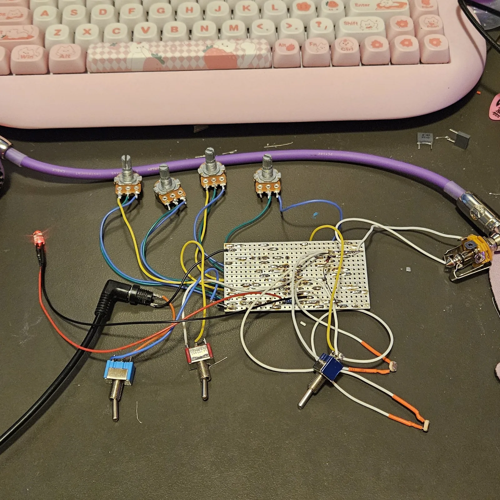The finished board on a dark desk mat, wired but not yet boxed. A rectangular strip of perfboard sits in the middle with its solder side up, its rows of holes joined by bright blobs of solder. Four potentiometers with bare knurled shafts and no knobs lie above it on blue, yellow and teal wires, and three toggle switches with blue and red bodies lie below on white and orange wires. A red LED glows at the left on red and black leads, a right-angle DC power plug enters from the lower left, and a quarter-inch jack with a purple instrument cable sits at the right. A pink cat-themed keyboard fills the top of the frame.