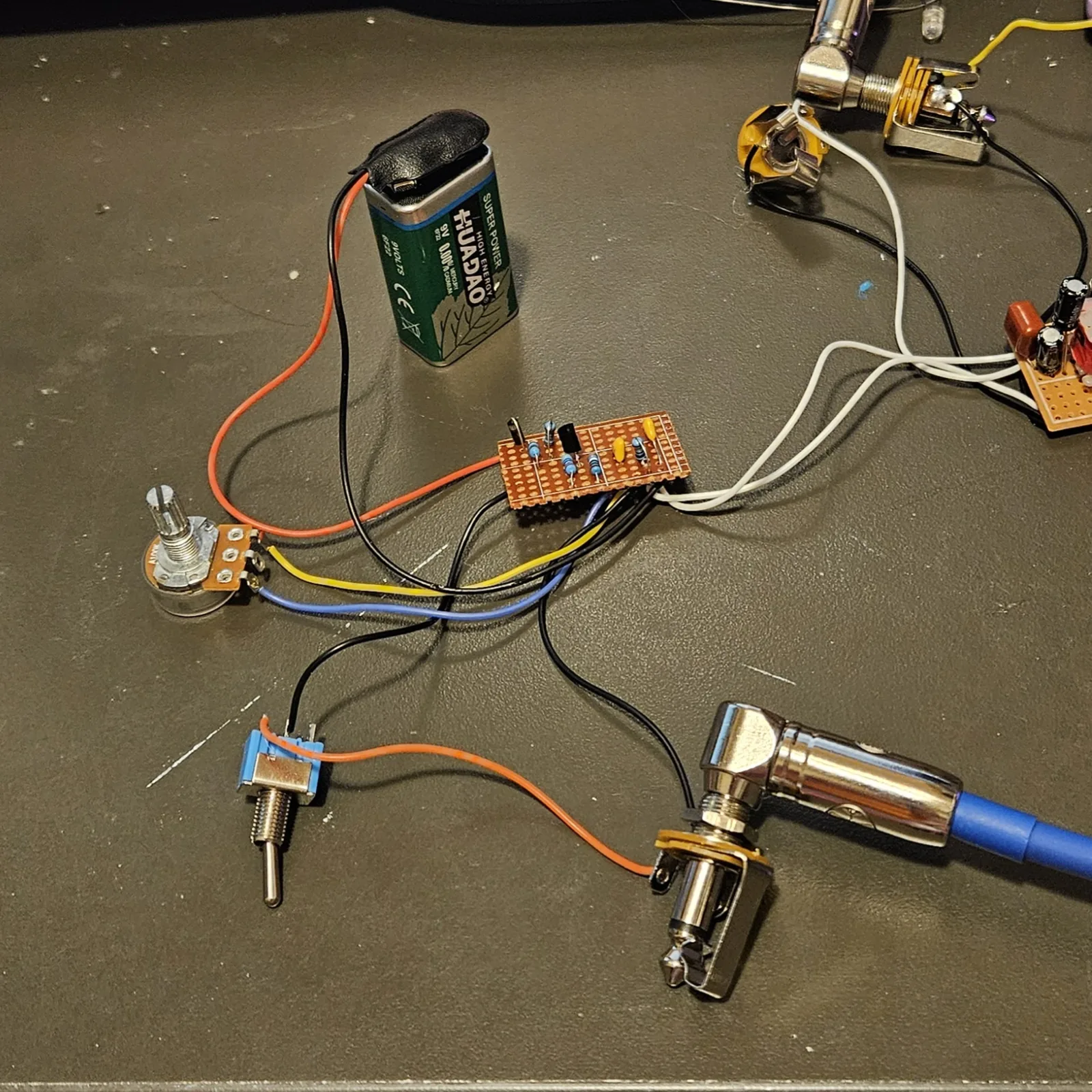 The Clean Boost built on a small piece of stripboard, lying on a dark bench with its parts standing up and wires trailing off it: a 9 V battery standing behind it, a potentiometer and a small blue toggle switch on flying leads, and a 1/4 inch jack with a blue right-angle plug in it. At the top right another jack and the ring modulator board with its two yellow transformers are wired to it.